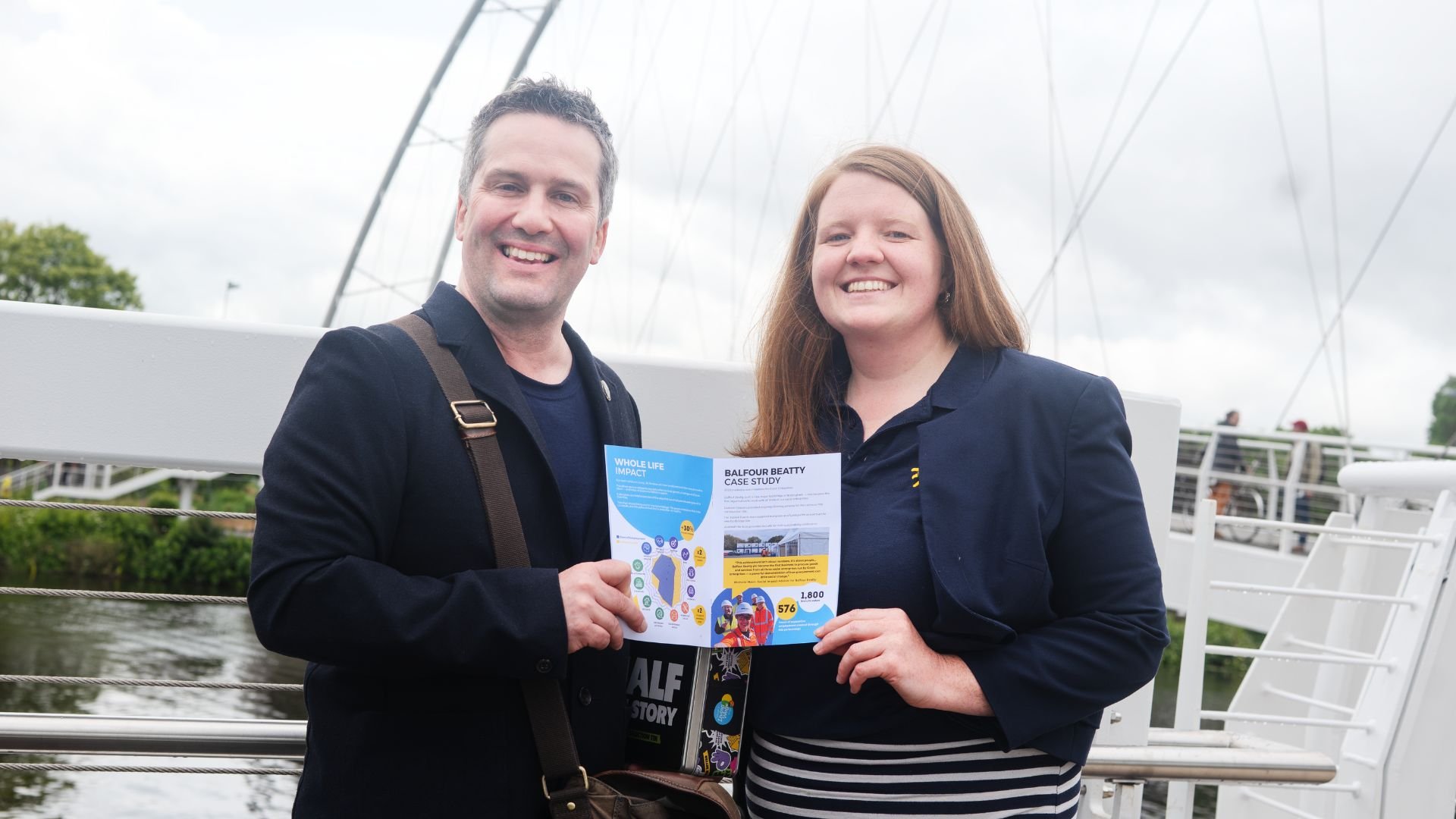 A man and woman smiling and holding a brochure together outdoors near a body of water with a bridge and people in the background.