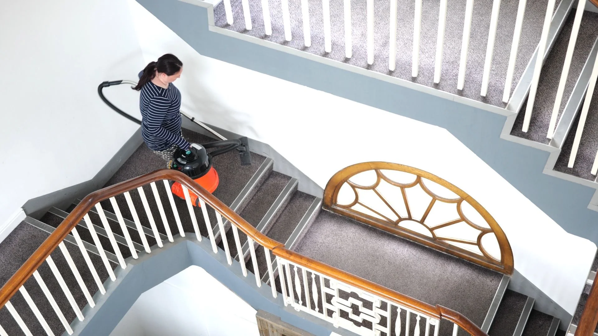 A woman vacuuming a carpeted staircase in a residential building, seen from above, with a wooden handrail and decorative wooden window grille.