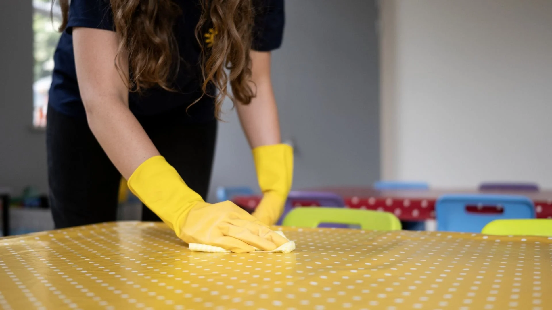 Person with long hair wearing yellow gloves cleaning a yellow polka dot tablecloth on a table with colorful chairs in the background.