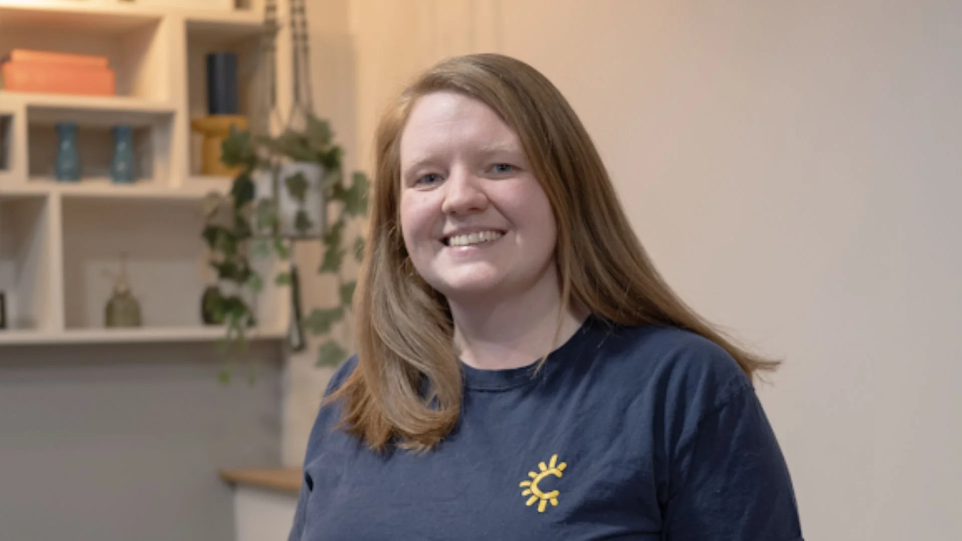 A woman with long red hair smiling at the camera, wearing a navy blue shirt with a yellow sun logo on the chest, in a room with a bookshelf and houseplants in the background.