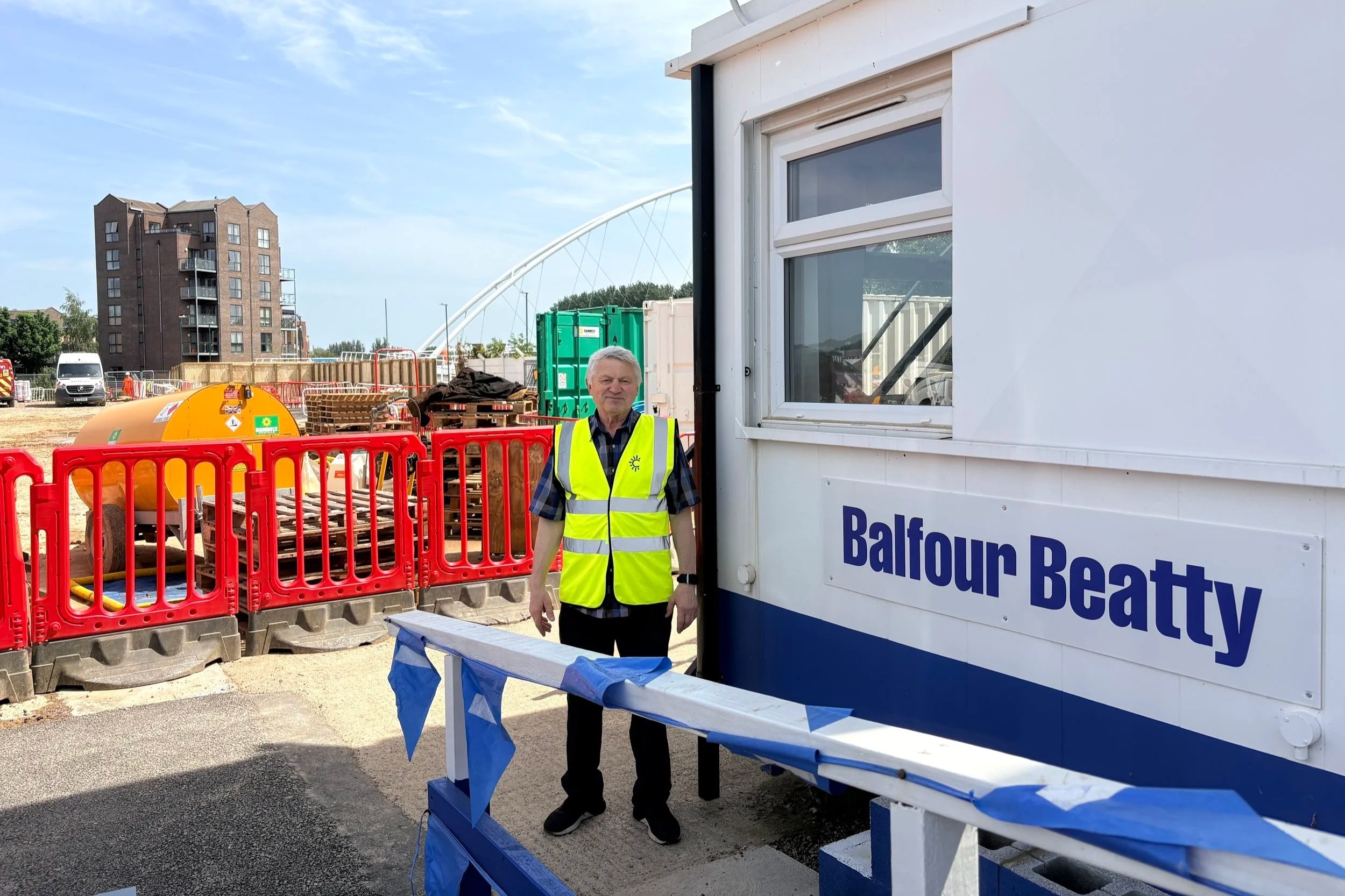 A man in a yellow high-visibility vest standing next to a portable battery station at a Balcour Beatty construction site. There are safety barriers, construction equipment, a white vehicle, and a building under construction in the background.