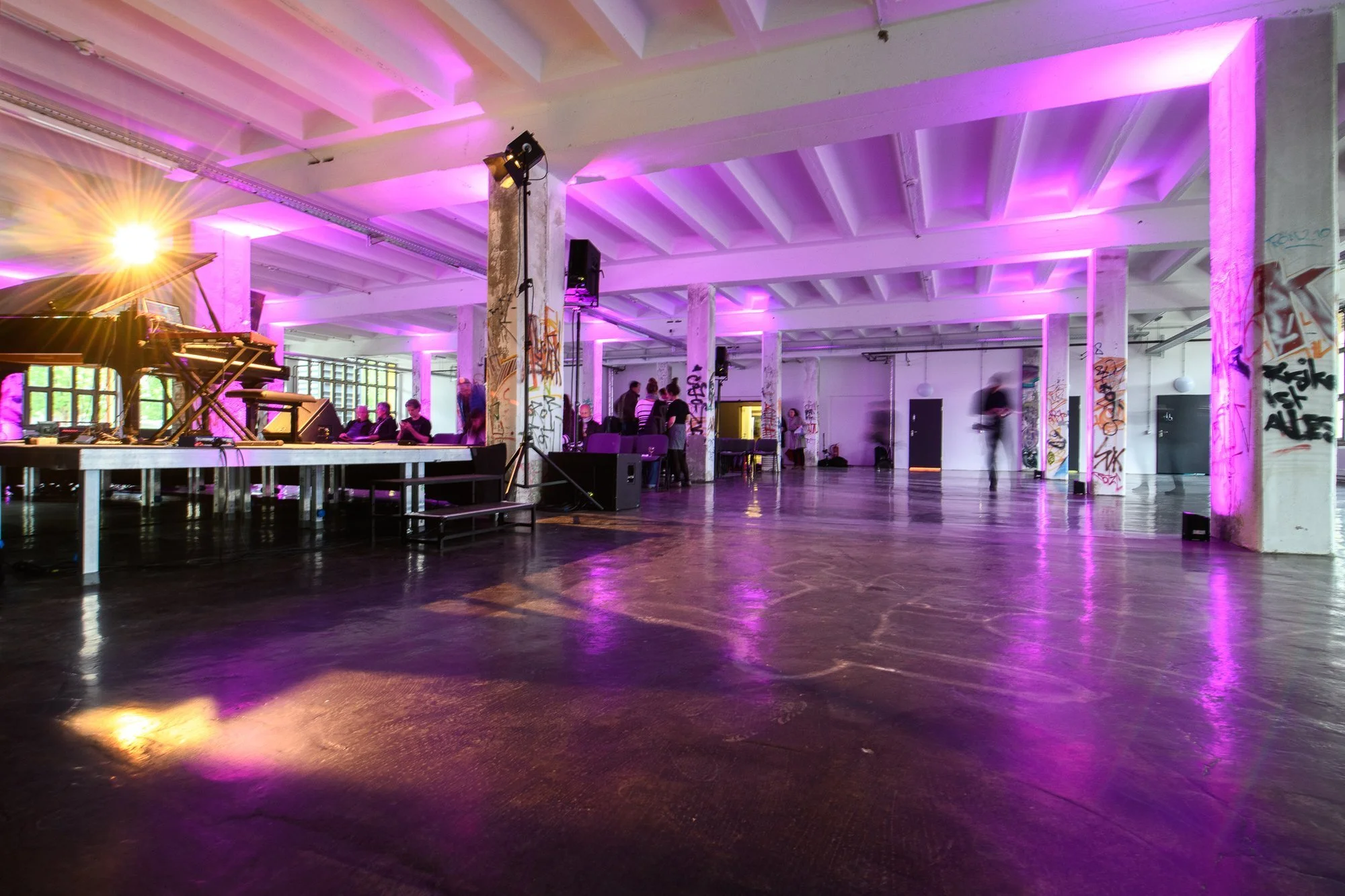 Innenraum einer Veranstaltungshalle mit Graffiti-Wänden, pinker Beleuchtung, einem Klavier und einigen Leuten, die sich unterhalten.