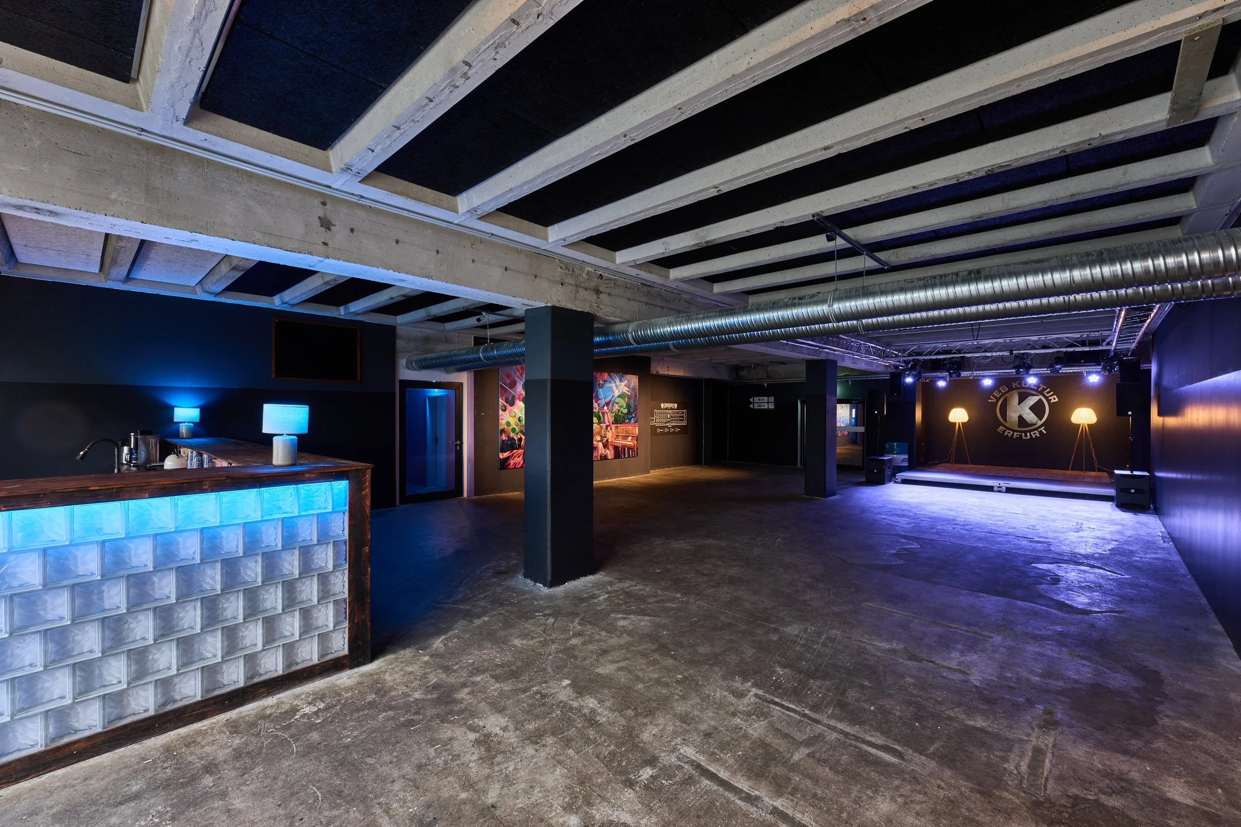Leeres Konzert- oder Veranstaltungsraum mit Bühne, schwarzer Wand mit Logo 'VEB Kultur', bunten Wandbildern, Bar mit blauen Lichtern, eleganten Steh-Lampen auf der Bühne, schwarzem Boden und sichtbaren Deckenstrukturen.