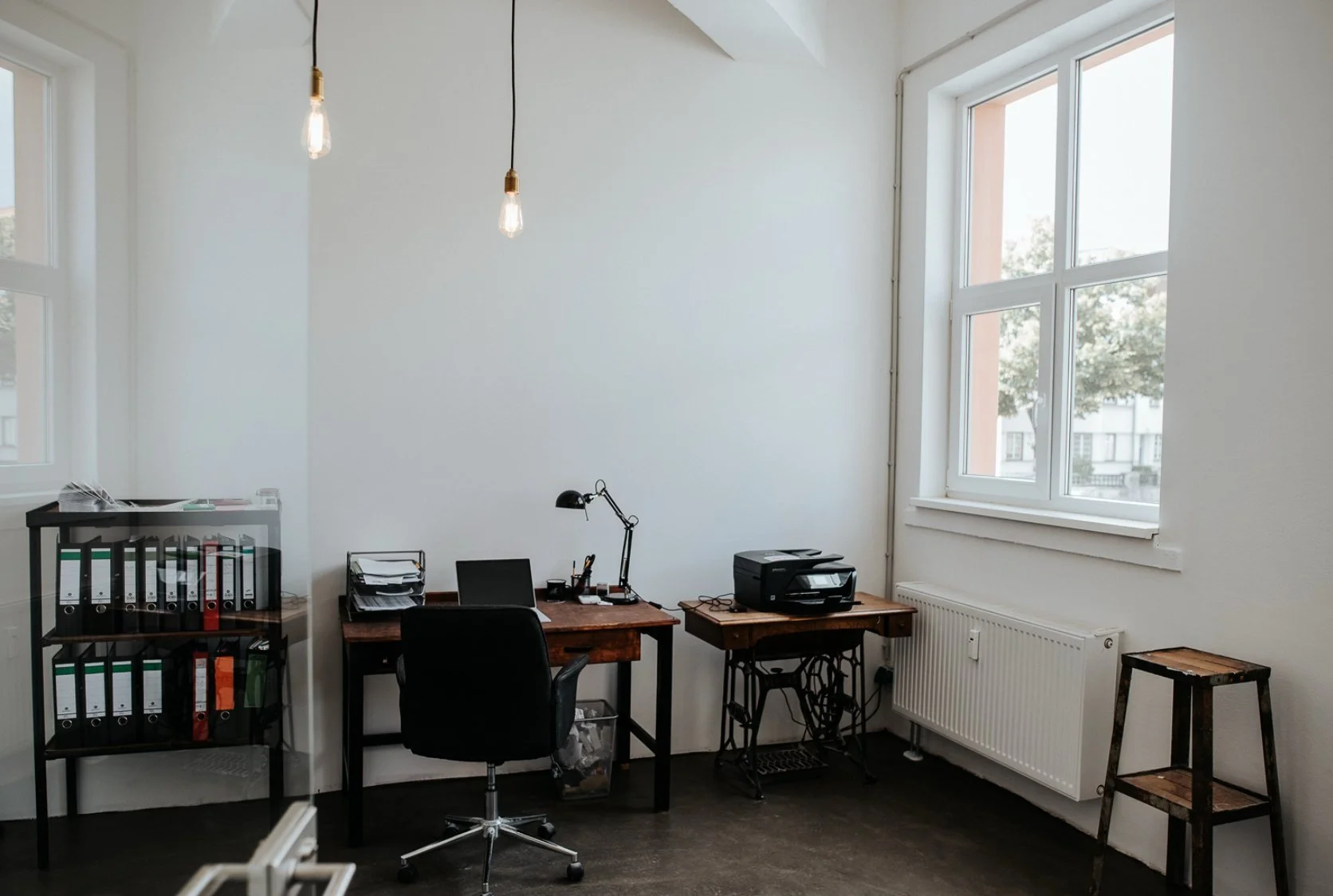Arbeitszimmer mit zwei Tischen, einem Bürostuhl, Aktenordner auf einem Regal, Drucker, Schreibtischlampe, große Fenster und eine Heizkörper