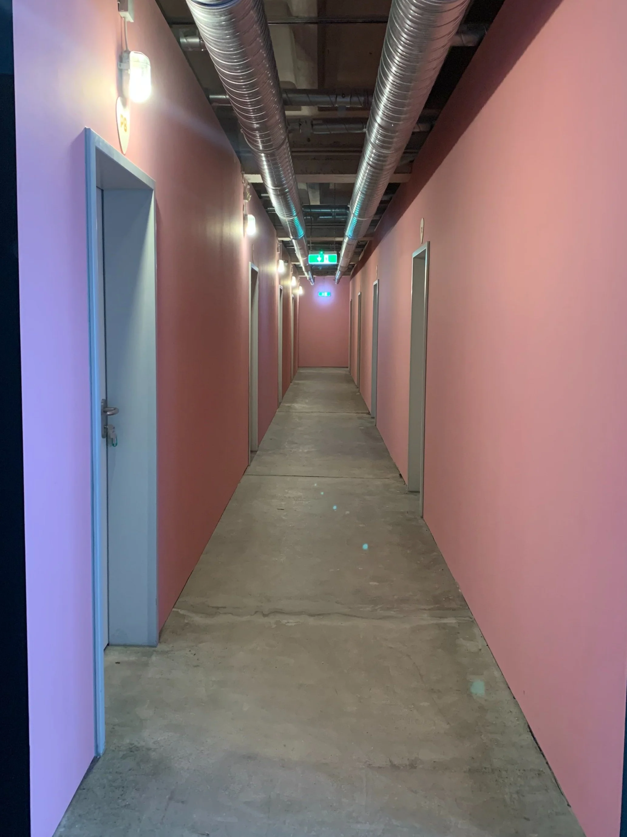 Ein langer Flur mit pinkfarbenen Wänden, mehreren Türen und Deckenventilatoren, sowie einer grünen Notausgangsschilderleuchte am Ende des Flurs.