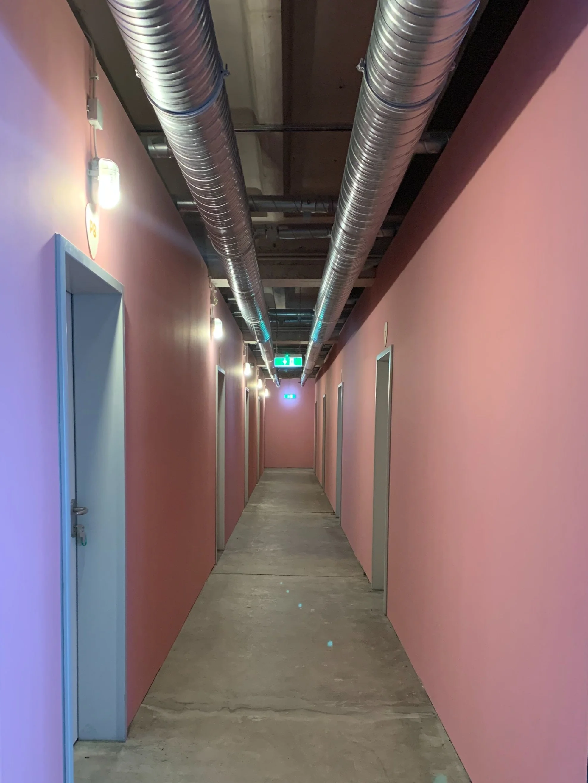 Langer Flur in einem Gebäude mit pinkfarbenen Wänden, mehreren Türen, error auch mit Türen und Rohren an der Decke.