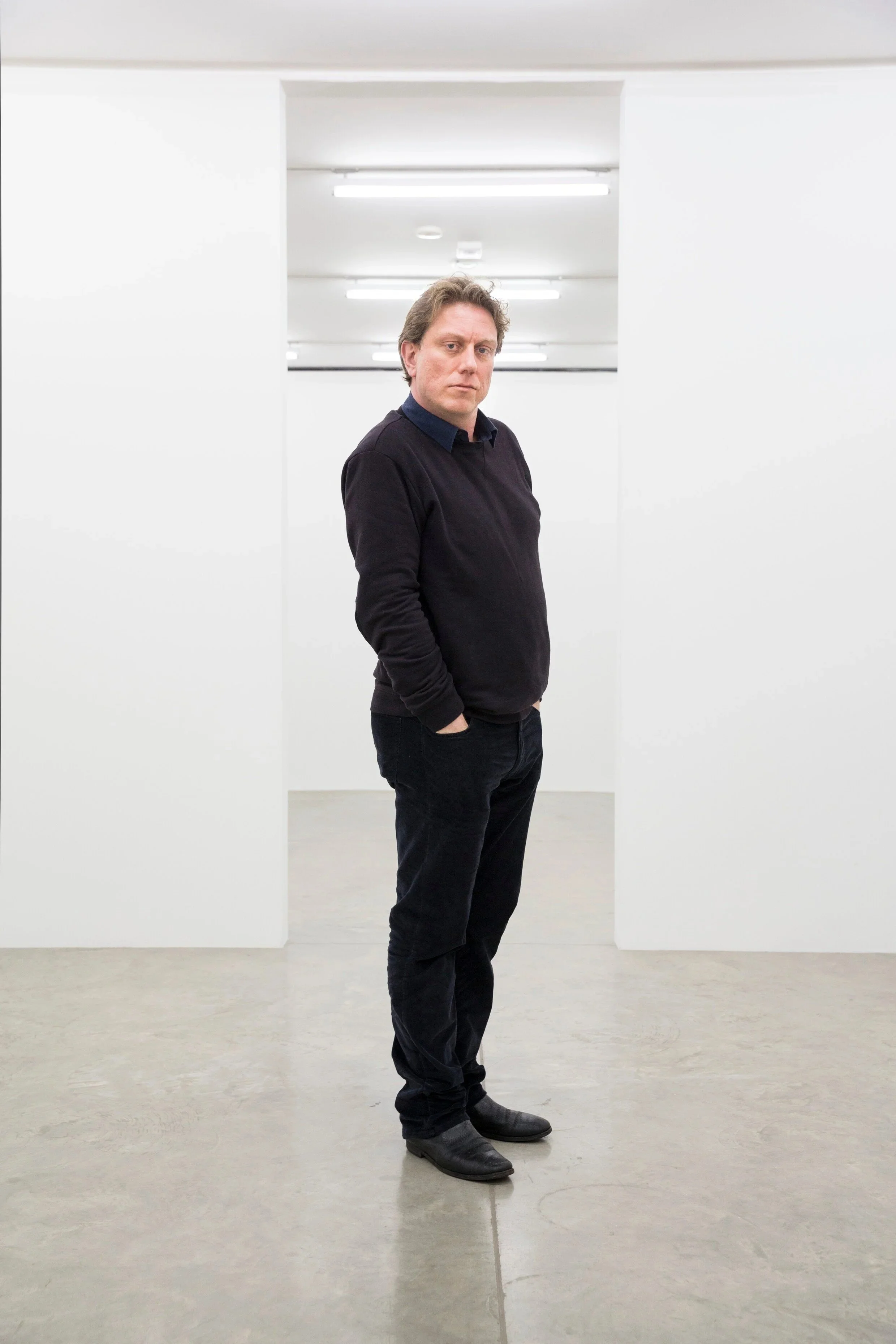A man standing in an all-white art gallery or museum with his hands in his pockets, wearing a black sweater, black pants, and black shoes, looking at the camera.