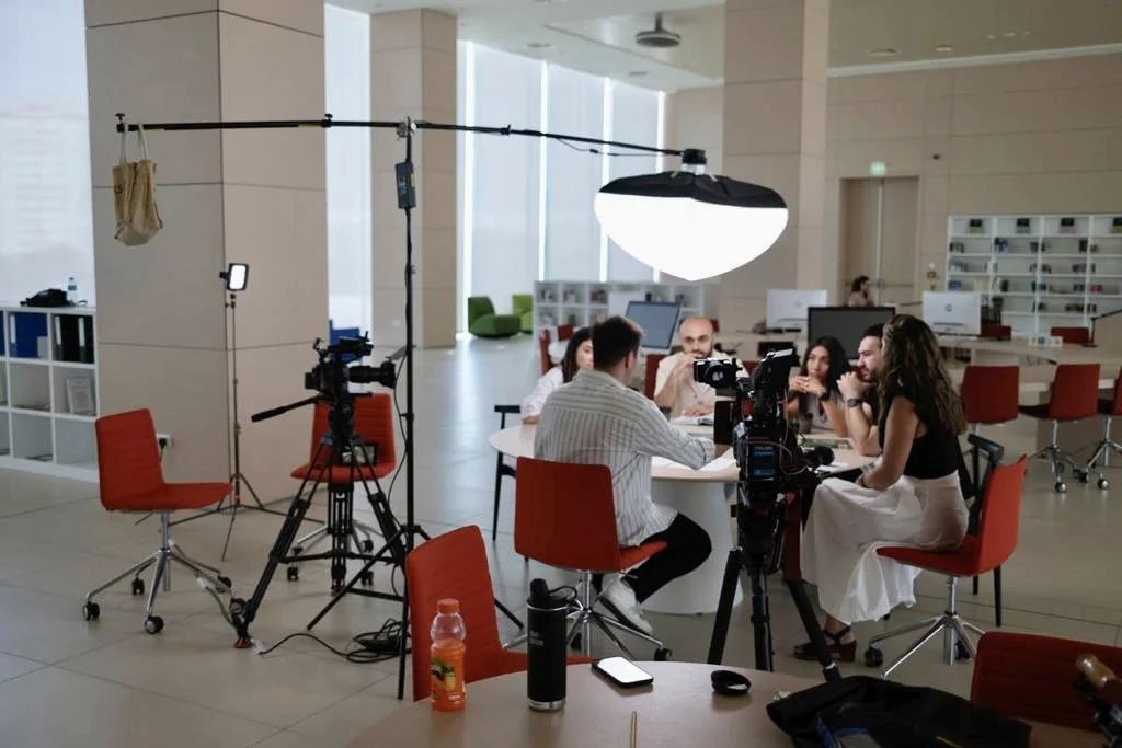 A group of people sitting around a table engaged in a discussion or interview, with professional lighting and video equipment set up for filming in a modern office space.