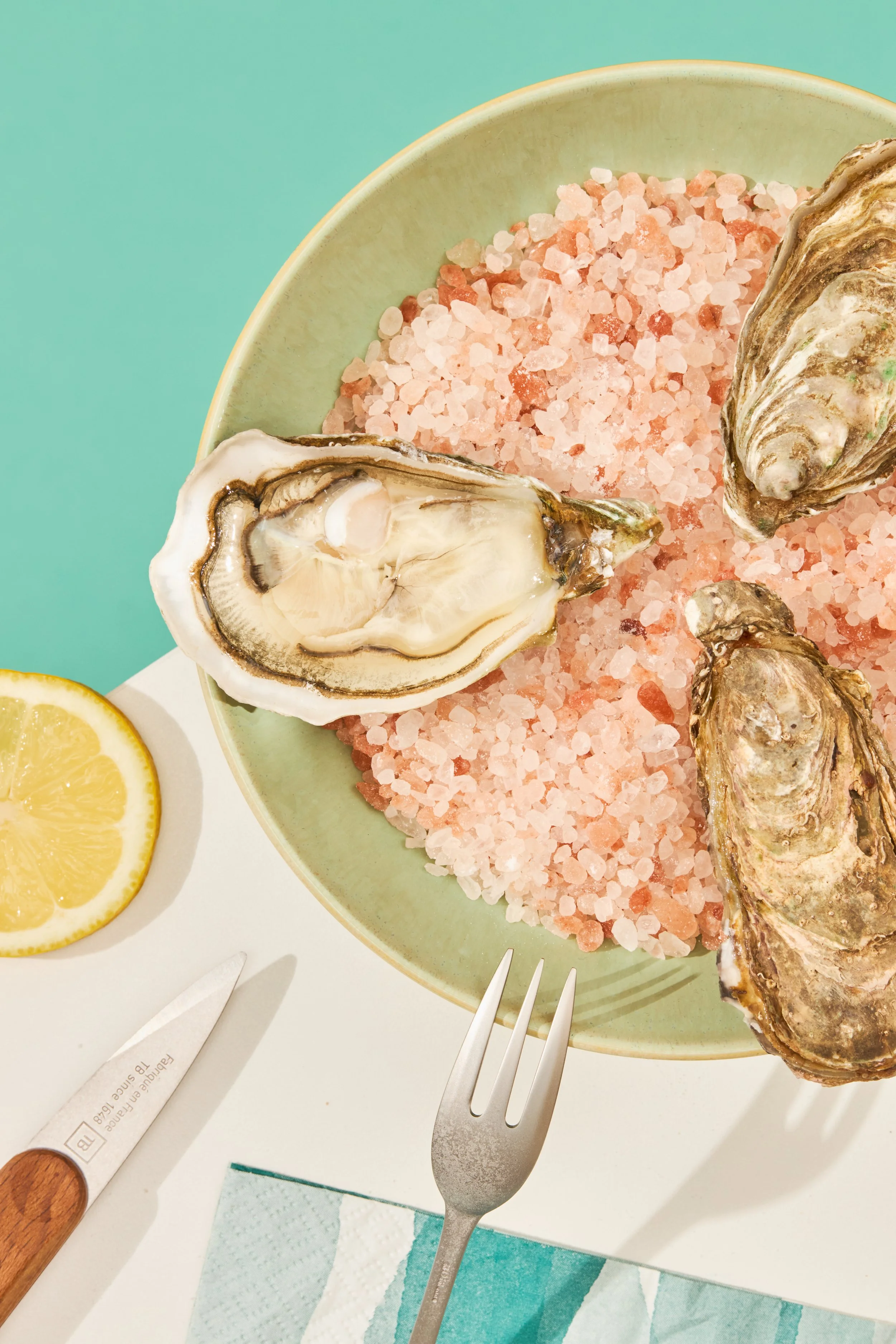 Une assiette verte contenant des huîtres et du sel rose de l'Himalaya, accompagnée de tranches de citron. Sur la table, il y a un couteau à huîtres, une fourchette et un torchon en papier.