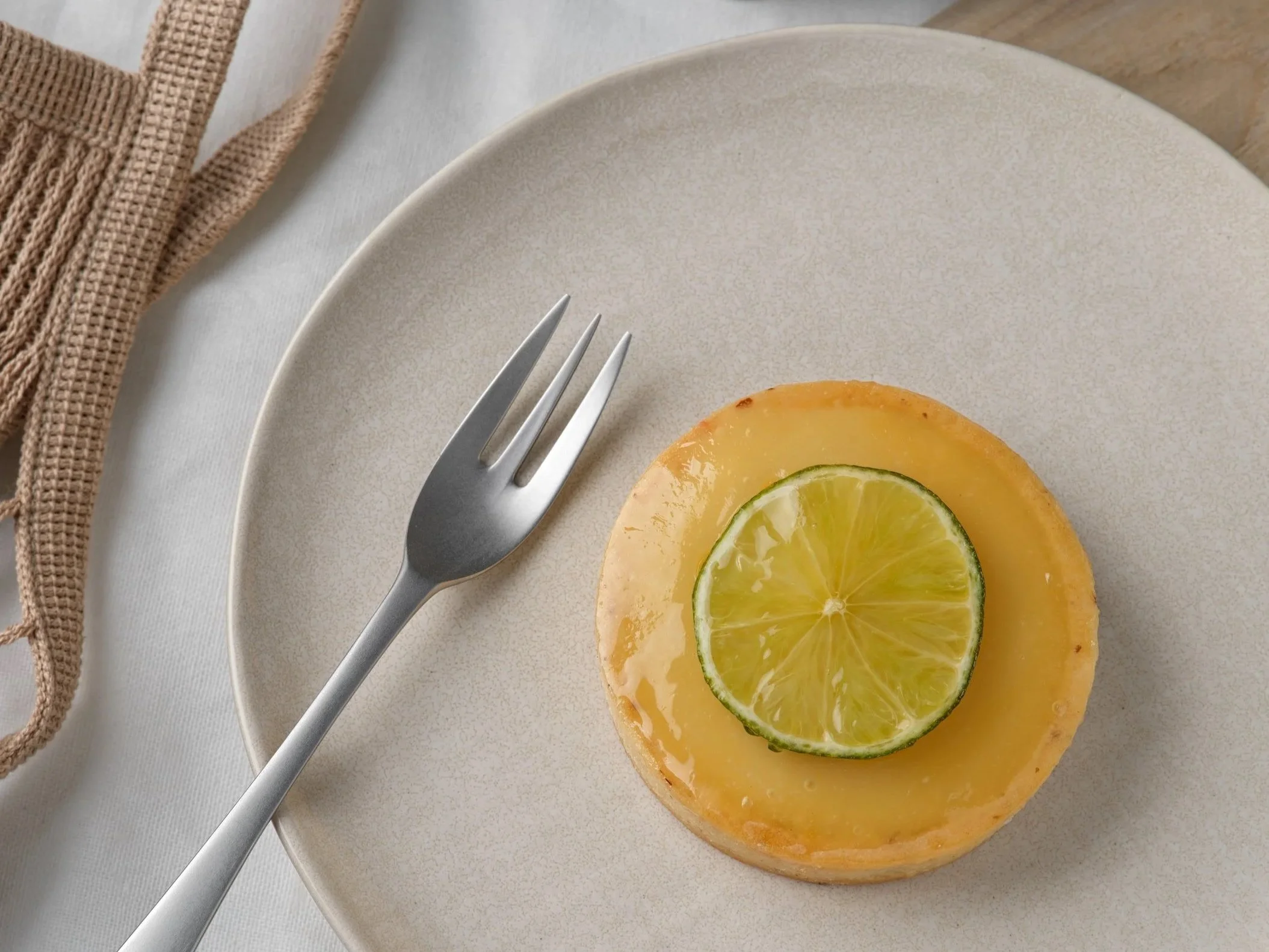 Une assiette beige avec une tartelette au citron vert. Une fourchette en métal est posé sur l'assiette, fond tissu et filet de course posé à coté.