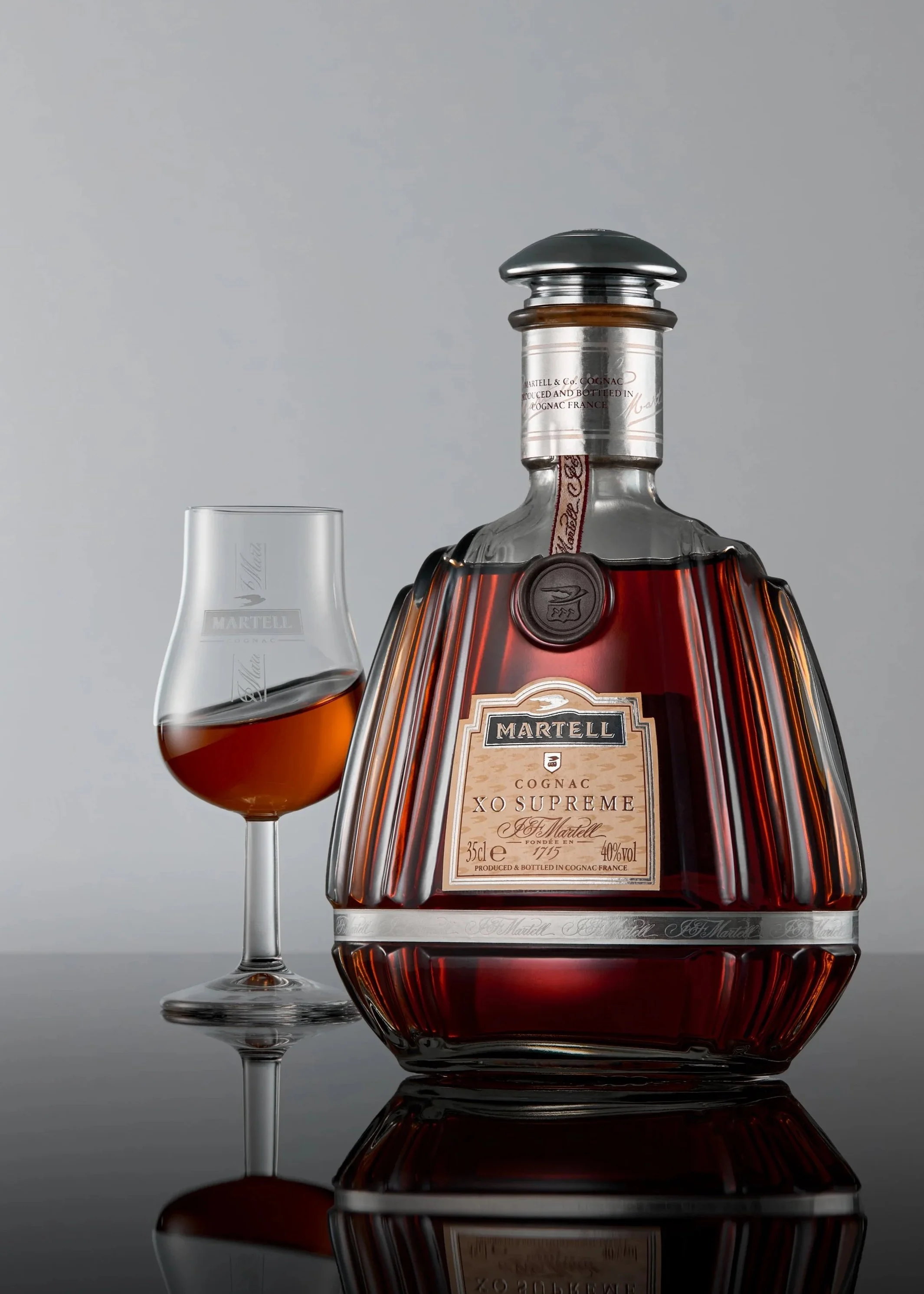 Une bouteille de cognac Martell XO Supreme avec un verre de cognac à côté, tous deux posés sur une surface réfléchissante grise.
