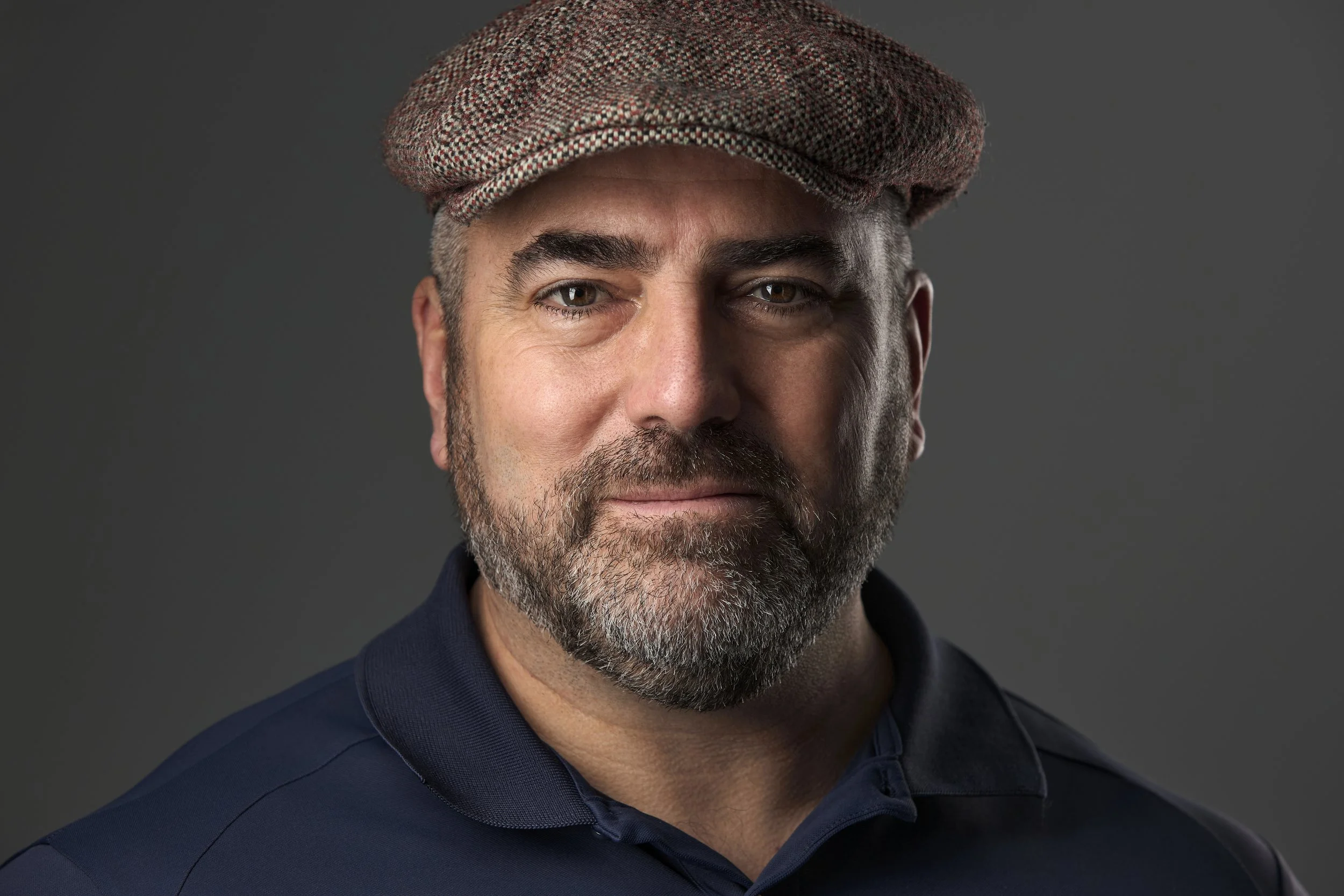 Portrait haut de gamme d'Homme avec barbe portant une casquette à carreaux de la marque stetson et un polo bleu foncé, regardant vers l'avant, fond gris