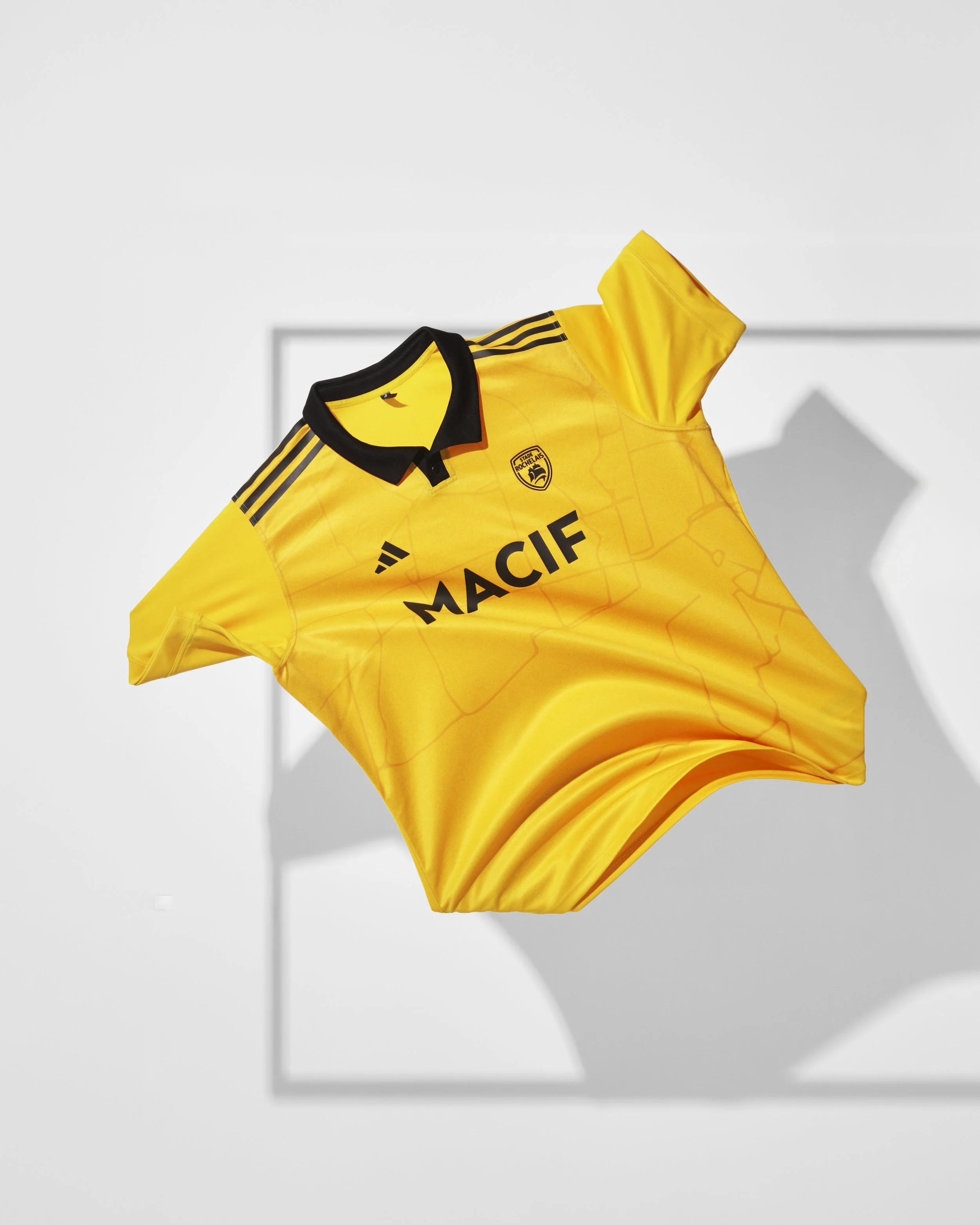 Maillot de rugby du stade rochelais jaune et noir avec logo du club et sponsor (macif), suspendu dans l'air, créant une ombre portée sur un fond blanc.
