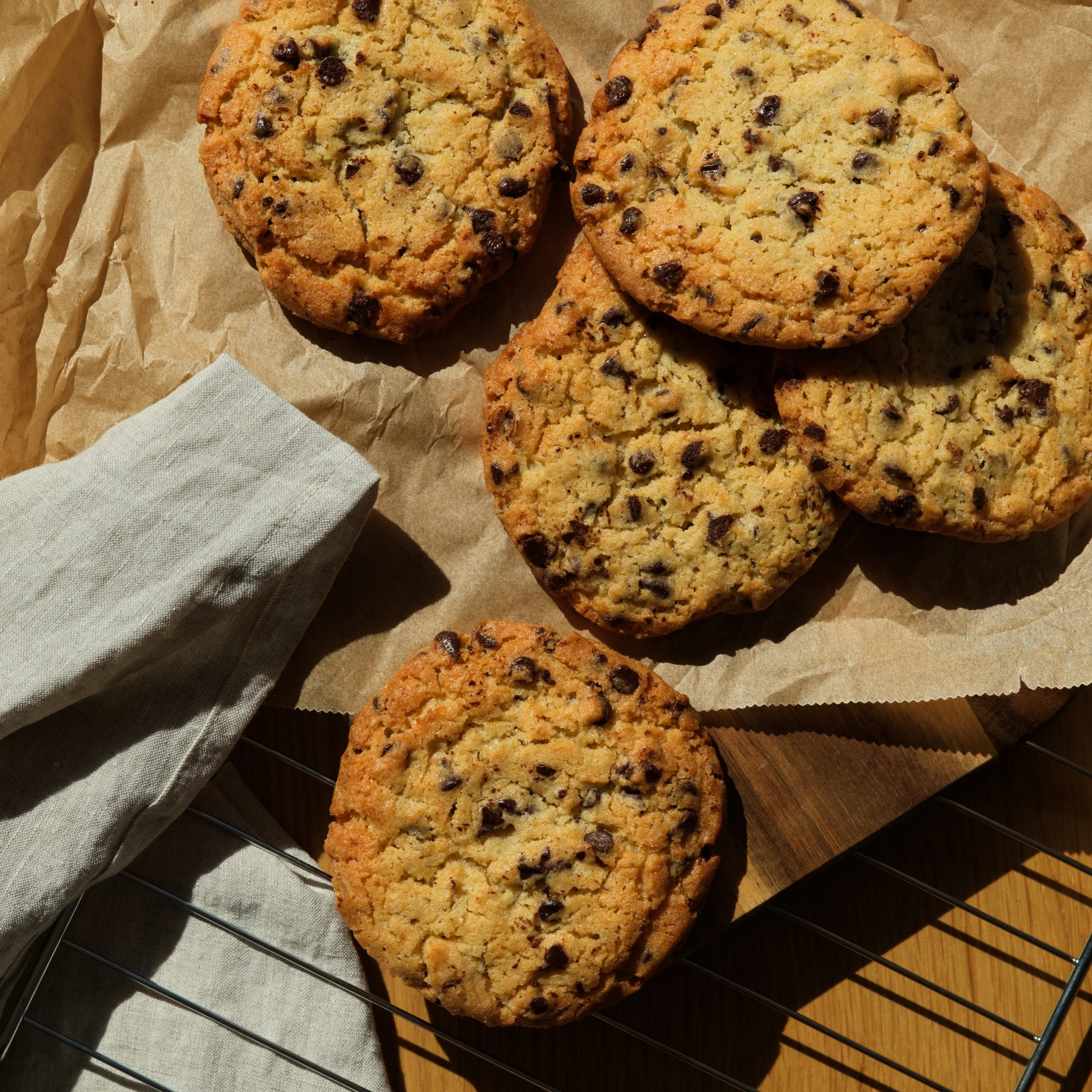 Cookies aux pépites de chocolat sur une grille en métal, avec une nappe beige en dessous et une table en bois à côté.