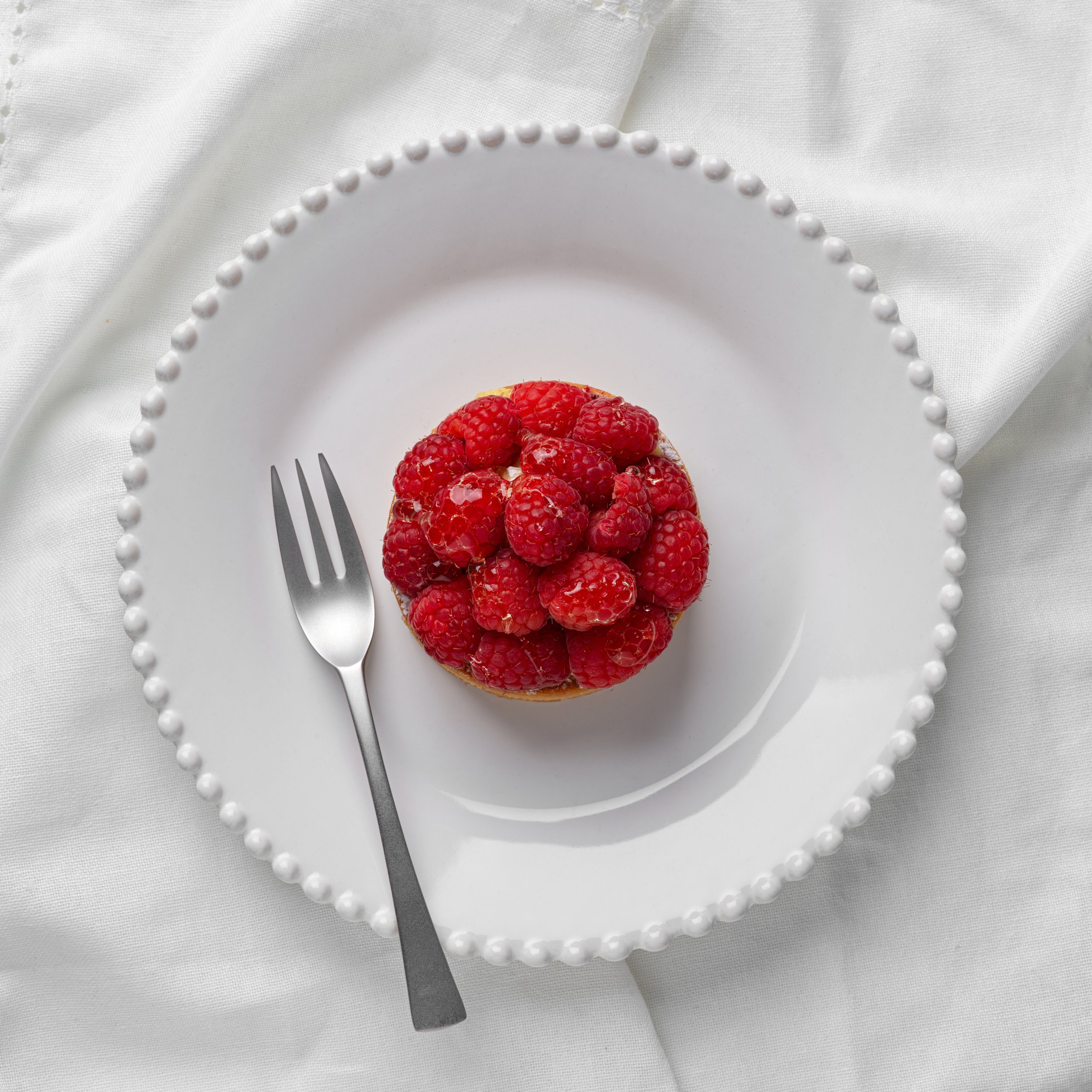Une tarte aux framboises rouges sur une assiette blanche, accompagnée d'une fourchette, sur un fond en tissu blanc.