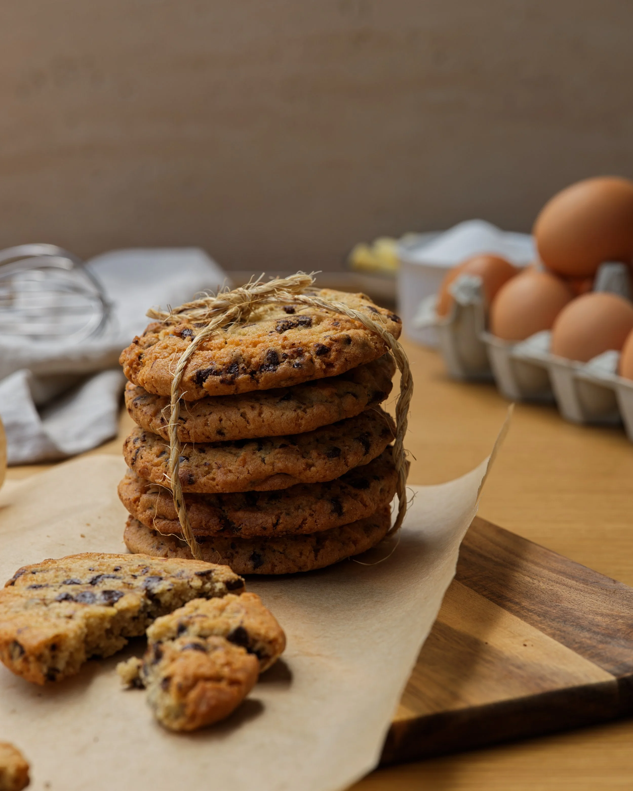 Une pile de biscuits cookies sur une feuille de papier cuisson. En arrière-plan, un boite à œufs et un fouet métallique.