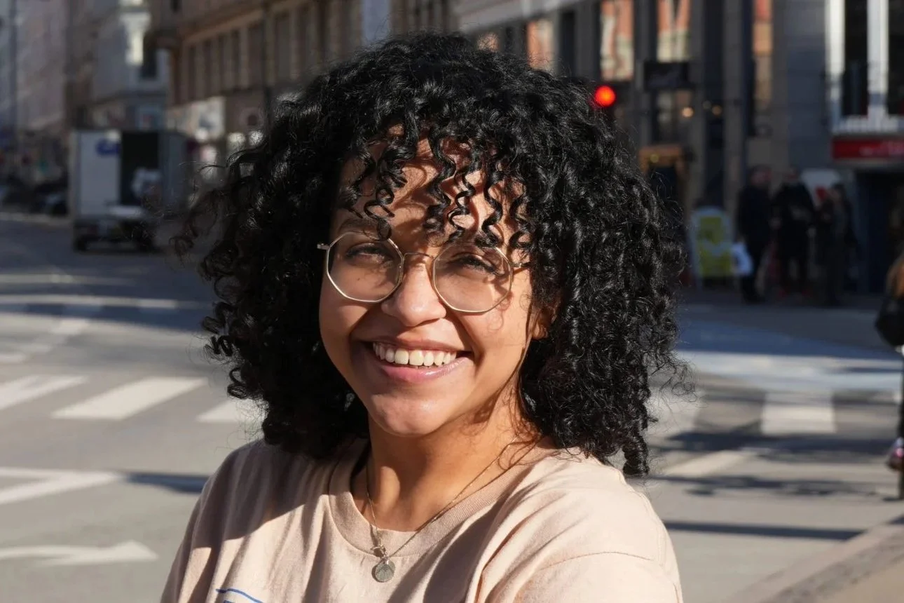 A woman with curly black hair, glasses, and a big smile standing on a city street.