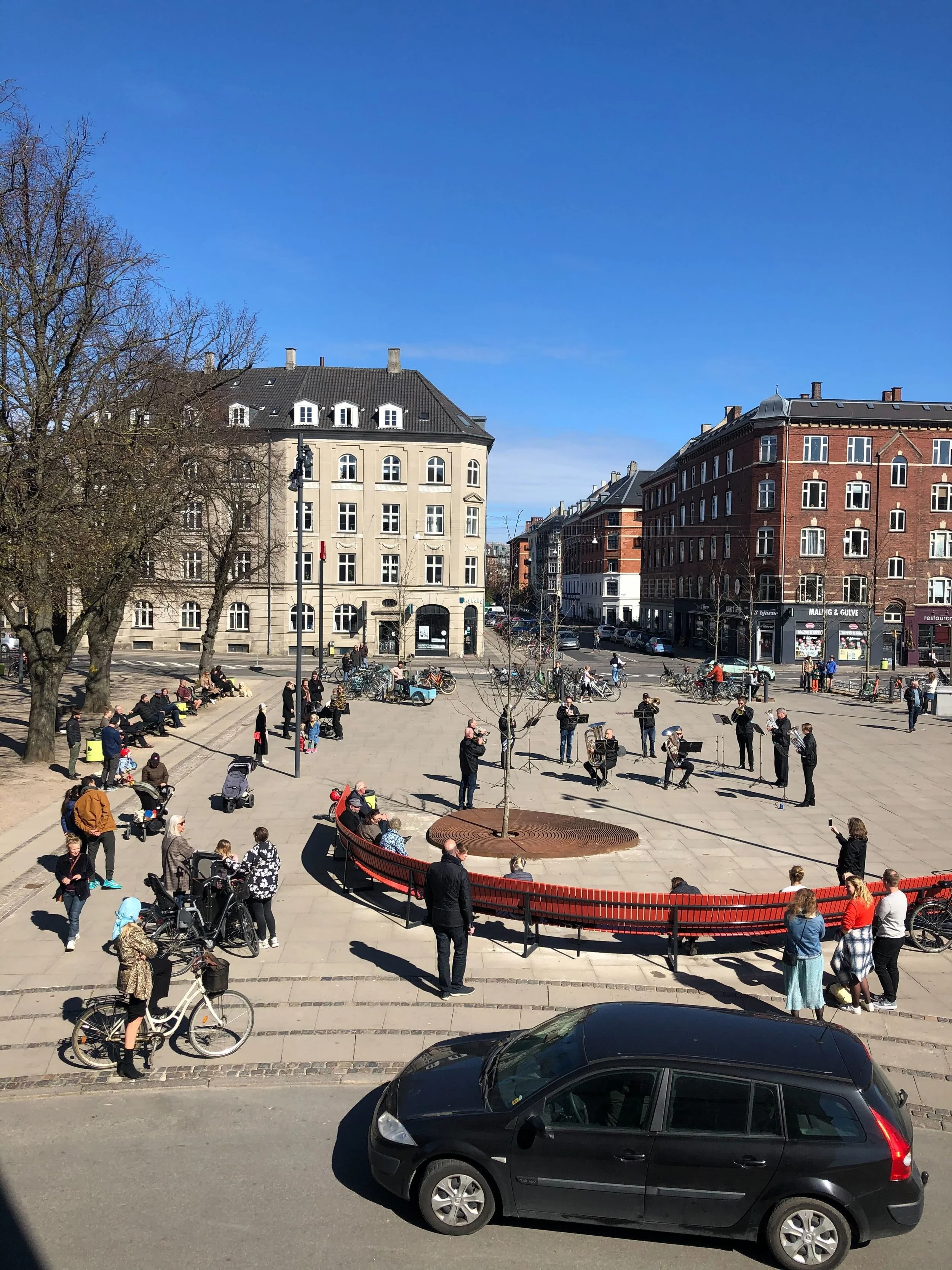 An outdoor scene of a city square with people sitting on benches, walking, and riding bicycles. A brass band is performing in the center, and surrounding buildings are visible under a clear blue sky.