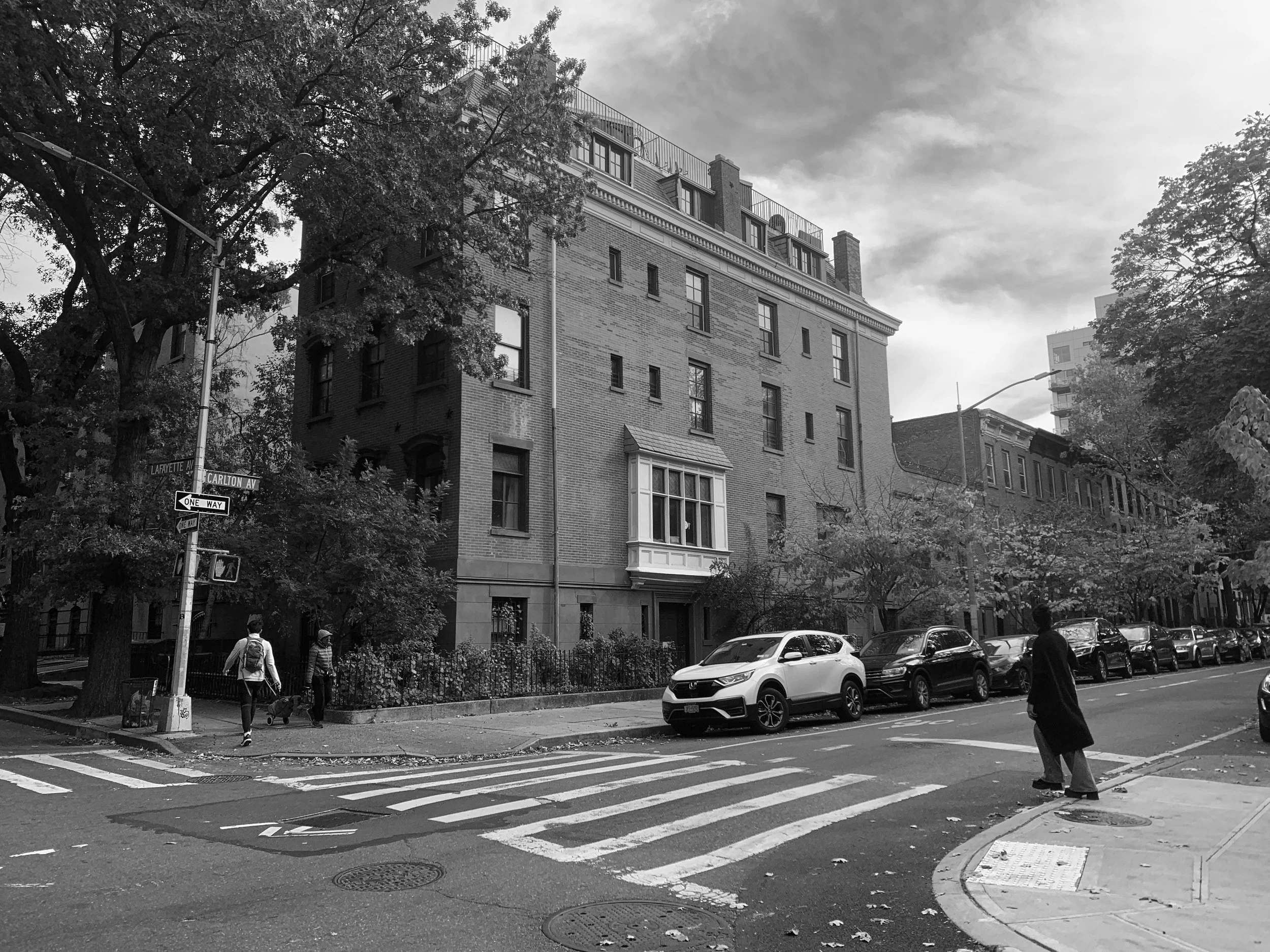 A black and white photo of a city street with a brick apartment building, trees, parked cars, and a crosswalk. Pedestrians are walking on the sidewalk.