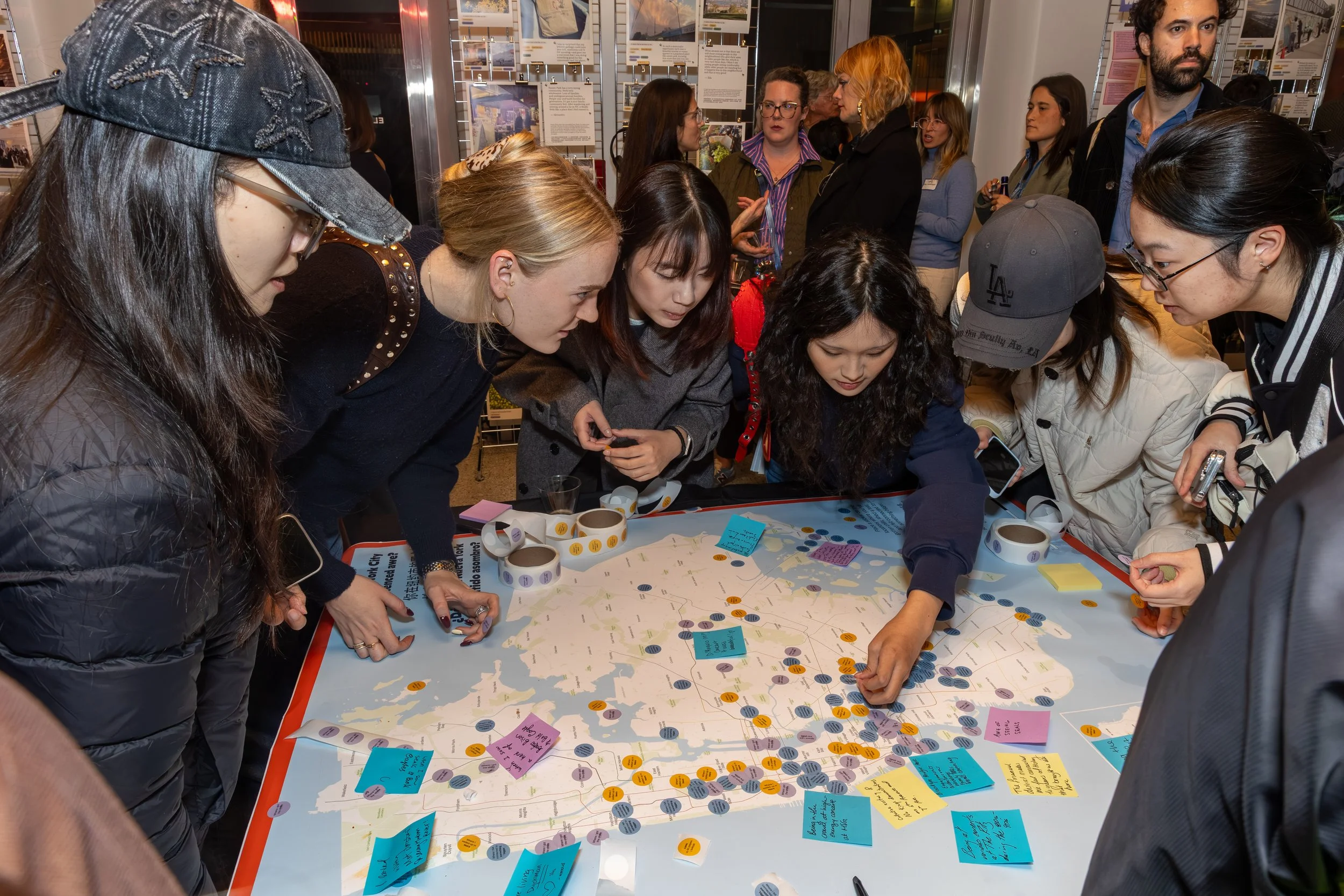 People gathered around a large map on a table, examining and discussing it. Some individuals are writing notes on sticky notes and using their phones. The background features display boards with information and photographs, indicating an indoor event or exhibit.