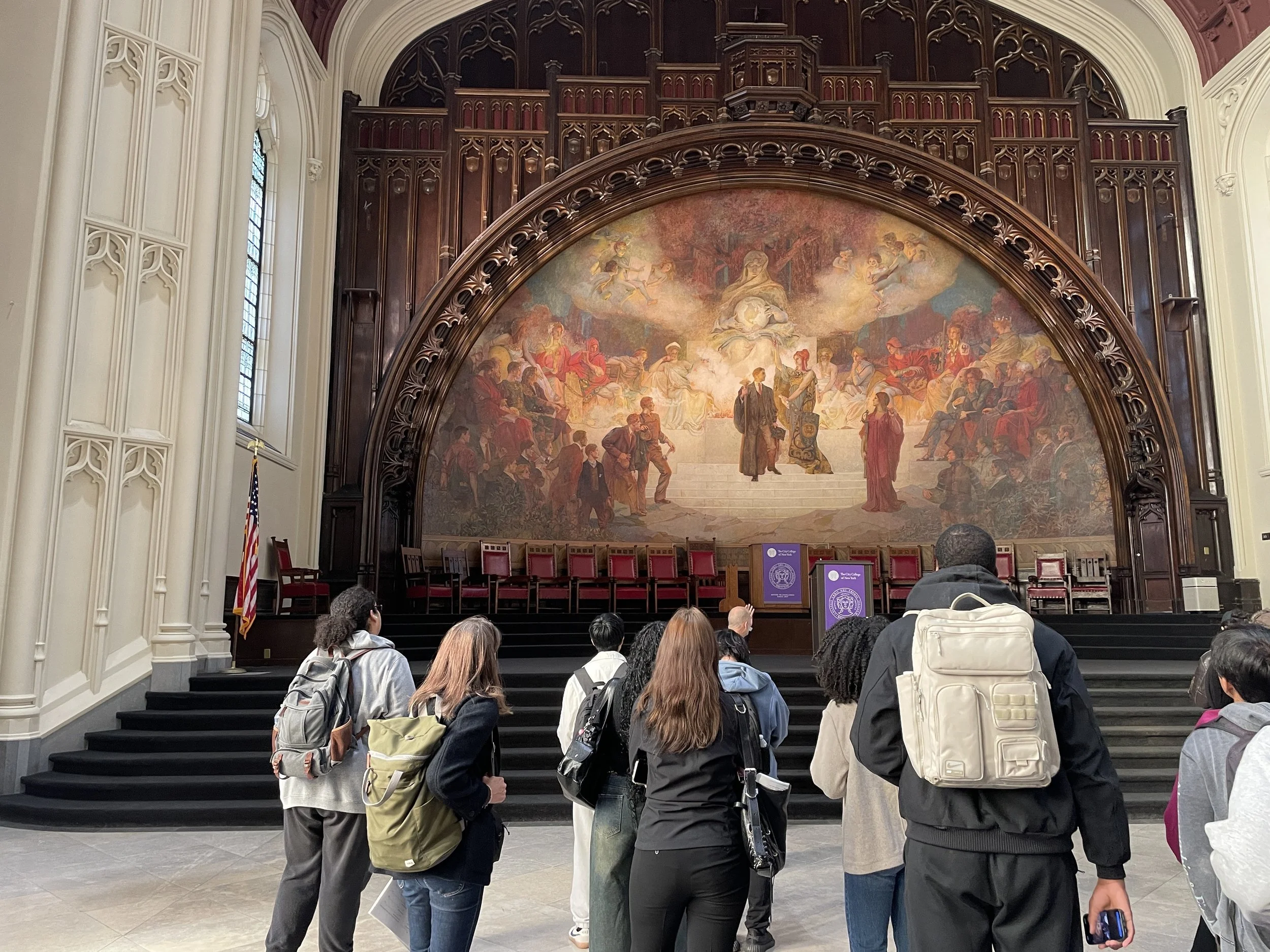 People standing in line in front of a large ornate stage with a mural, inside a church.