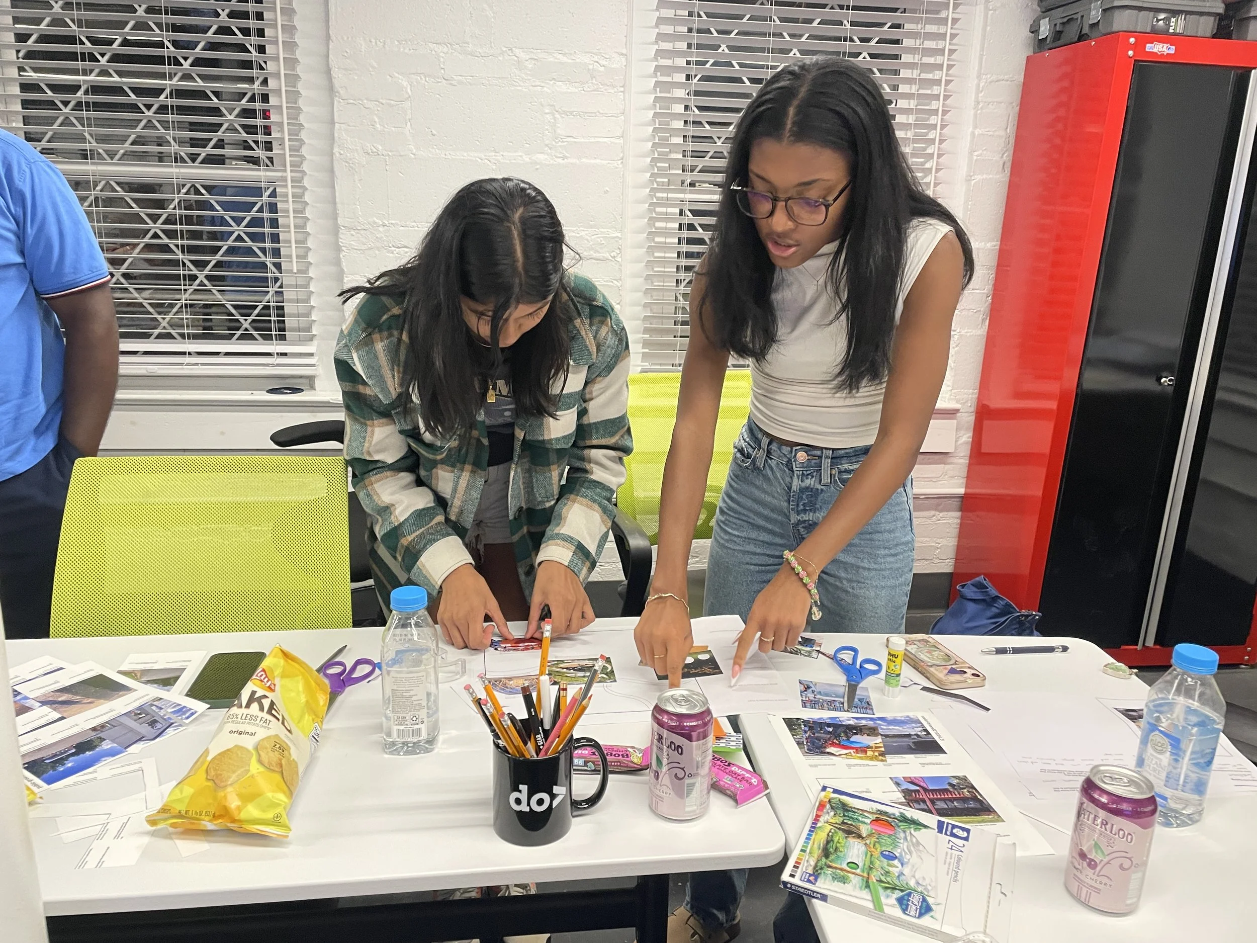 Two women engaged in a creative activity at a table covered with papers, photos, craft supplies, bottle of water, snacks, and drinks. They are bending over the table, working on a project. The setting appears to be an indoor room with windows and blinds, and a red cabinet is visible in the background.