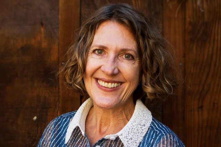 A woman with shoulder-length wavy hair smiling in front of a wooden background.