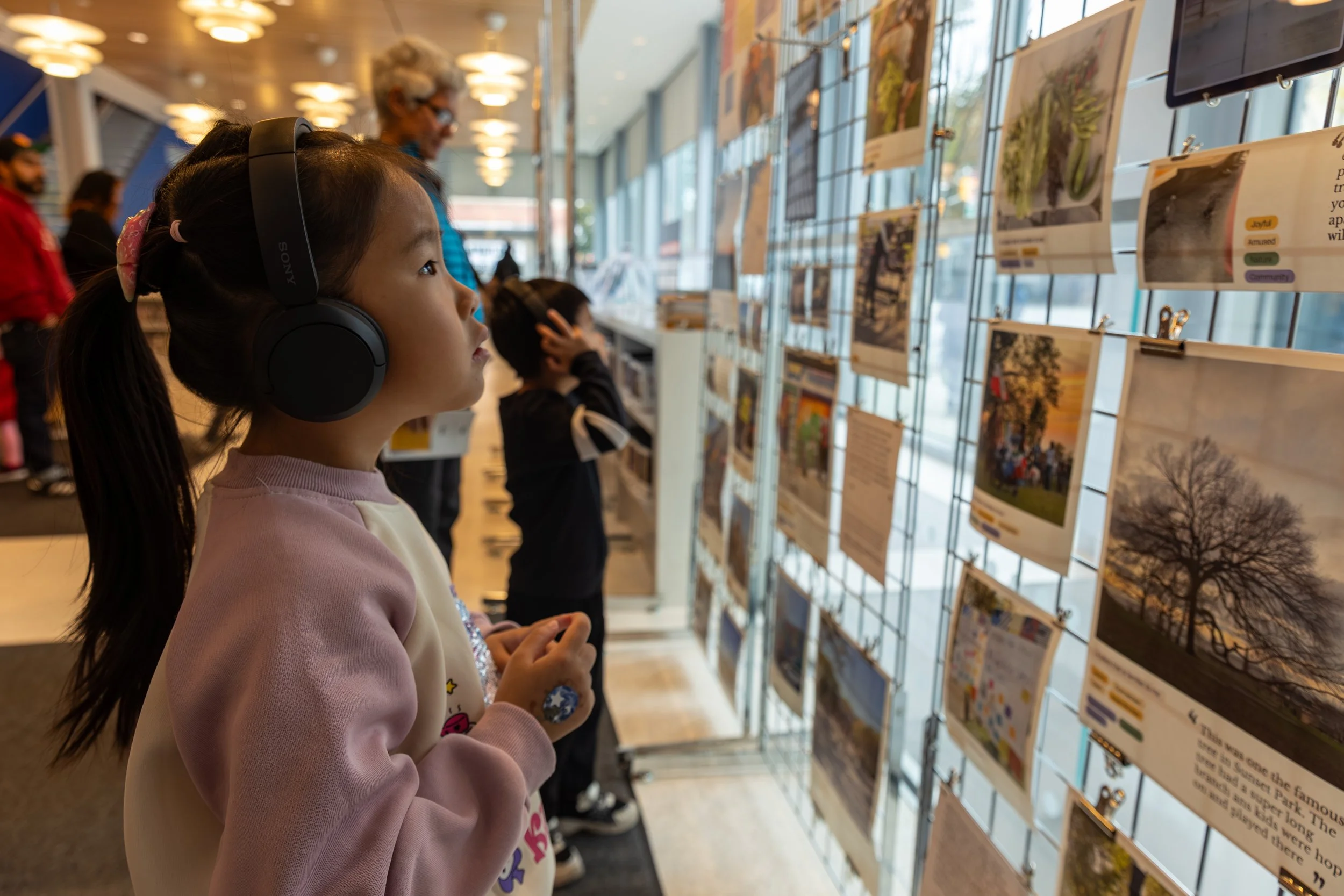 A young girl with dark hair wearing large black Sony headphones looking at artwork displayed in an exhibition.