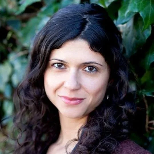 Close-up portrait of a woman with long, dark curly hair, smiling softly in front of green foliage.