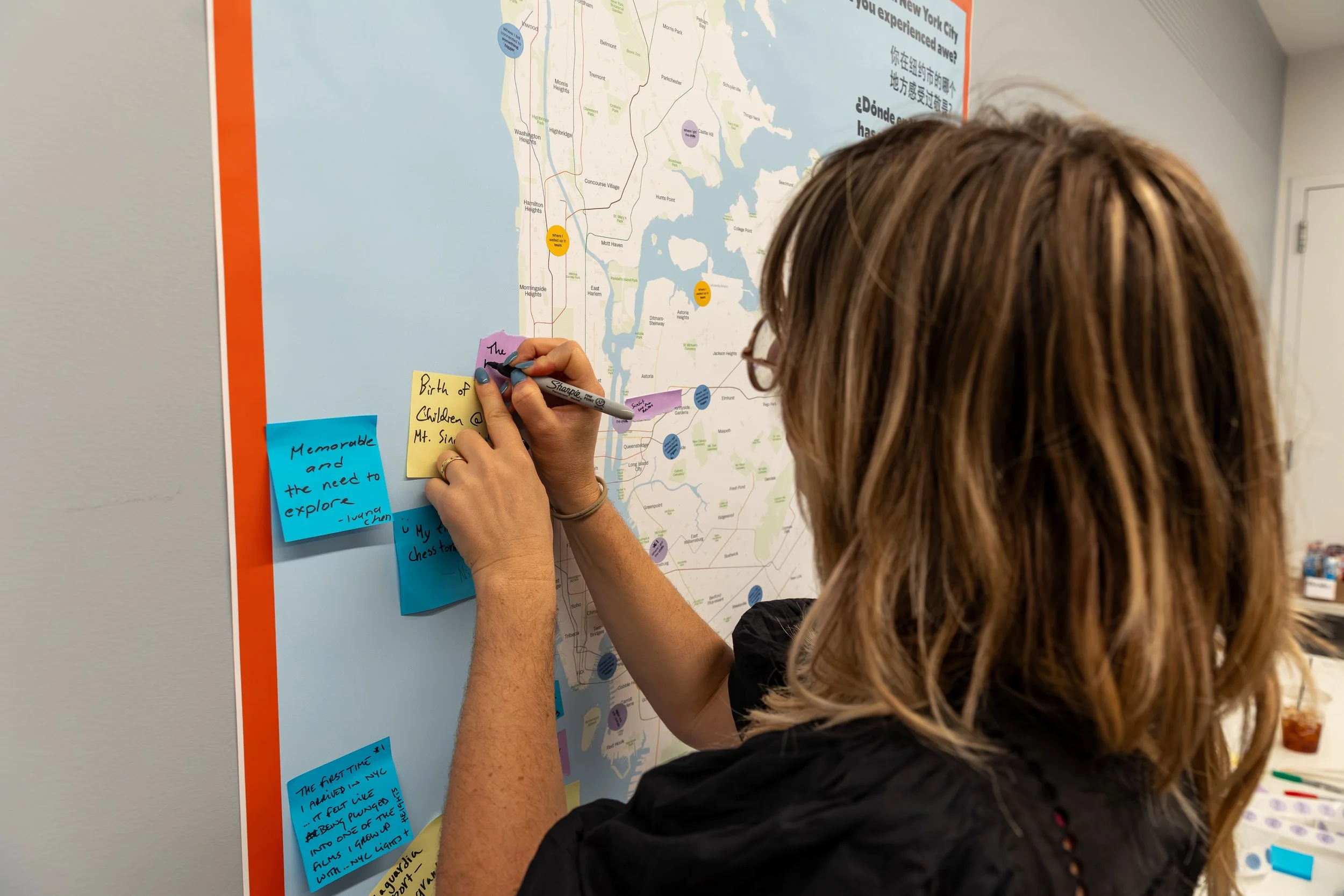 A person with light brown hair and glasses writing on sticky notes on a large map of New York City pinned to the wall. The map shows neighborhoods and colored dots indicating locations or points of interest, and the sticky notes contain handwritten notes.