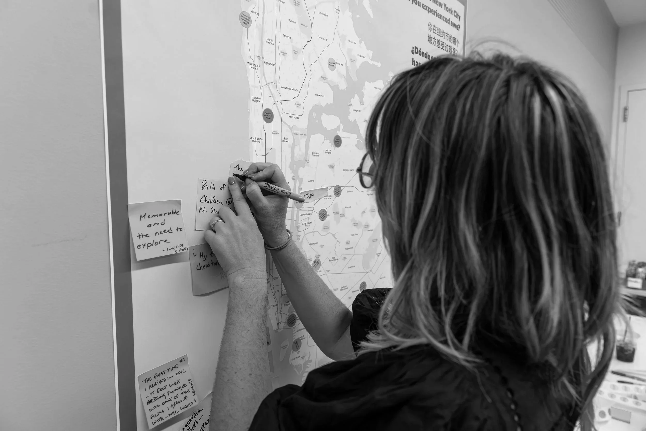 A person with shoulder-length hair and glasses writing notes on sticky notes attached to a large map of New York City, covered in both English and Chinese text.
