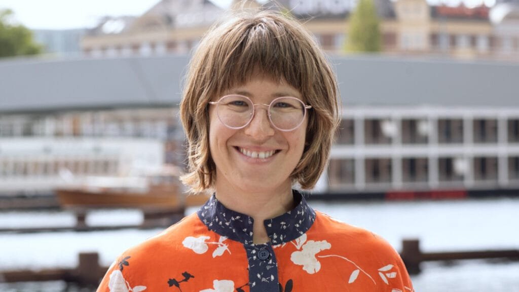 A smiling woman with glasses and short brown hair standing outside near a body of water with boats and buildings in the background.