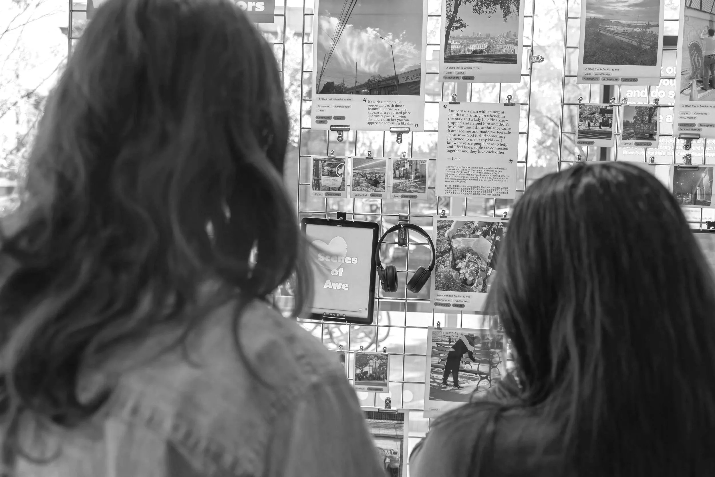Two people with long hair are looking at a display of photos and information on a wire grid. The display contains various images, texts, and labels such as "scenes of awe" and words like "joyful," "community," and "history." Some photos depict outdoor scenes, parks, and cityscapes.