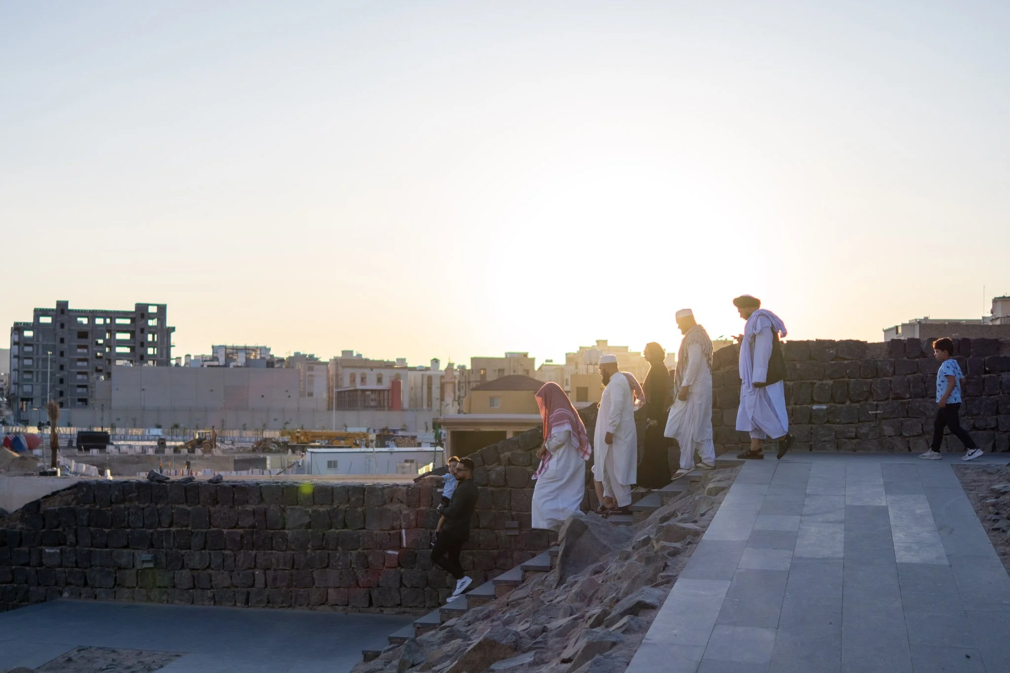 A group of men and boys dressed in traditional Middle Eastern clothing walking on a stone pathway near a stone wall during sunset, with urban buildings in the background.