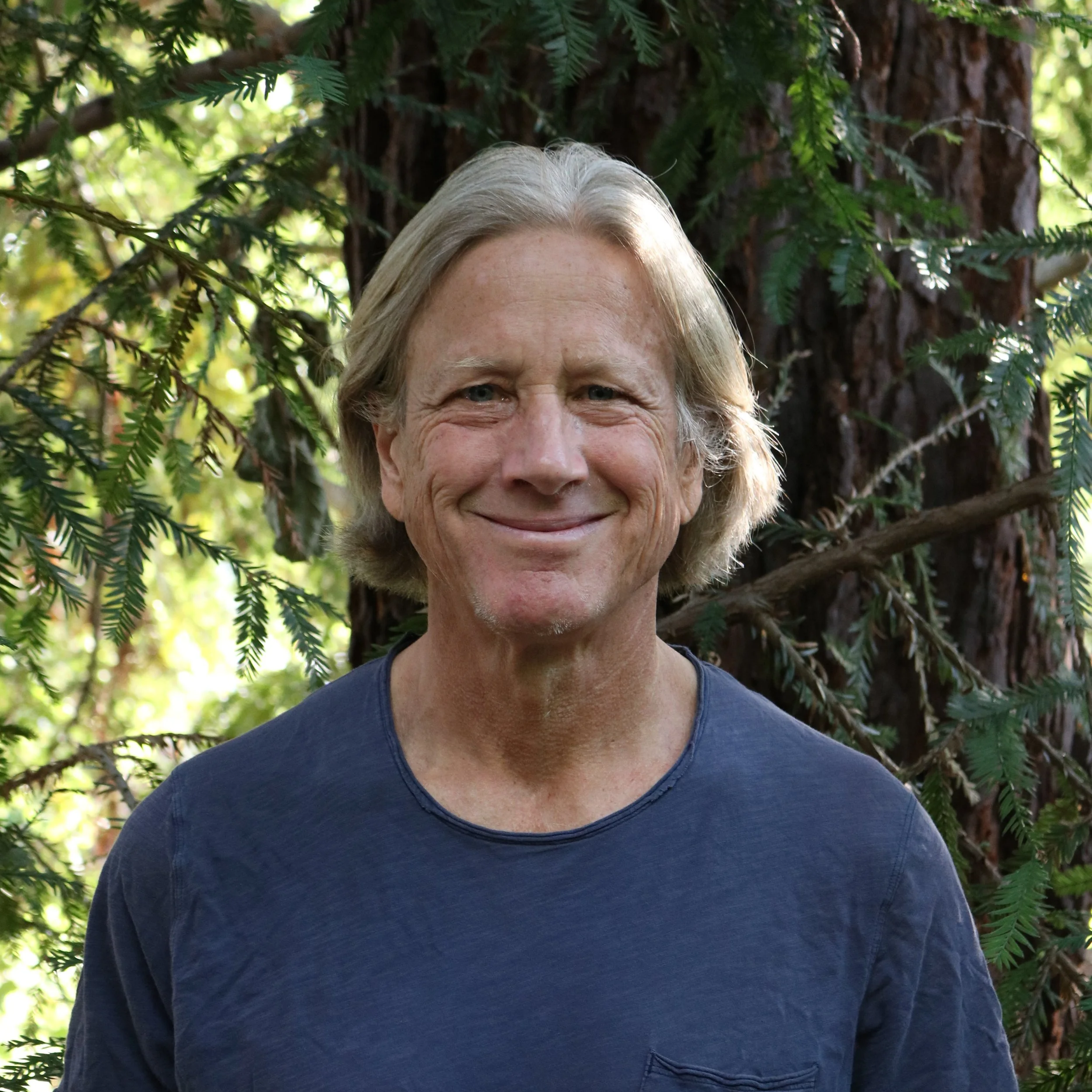 Close-up of an older man with gray, shoulder-length hair and blue eyes, smiling, outdoors surrounded by green pine tree branches and a large tree trunk in the background.