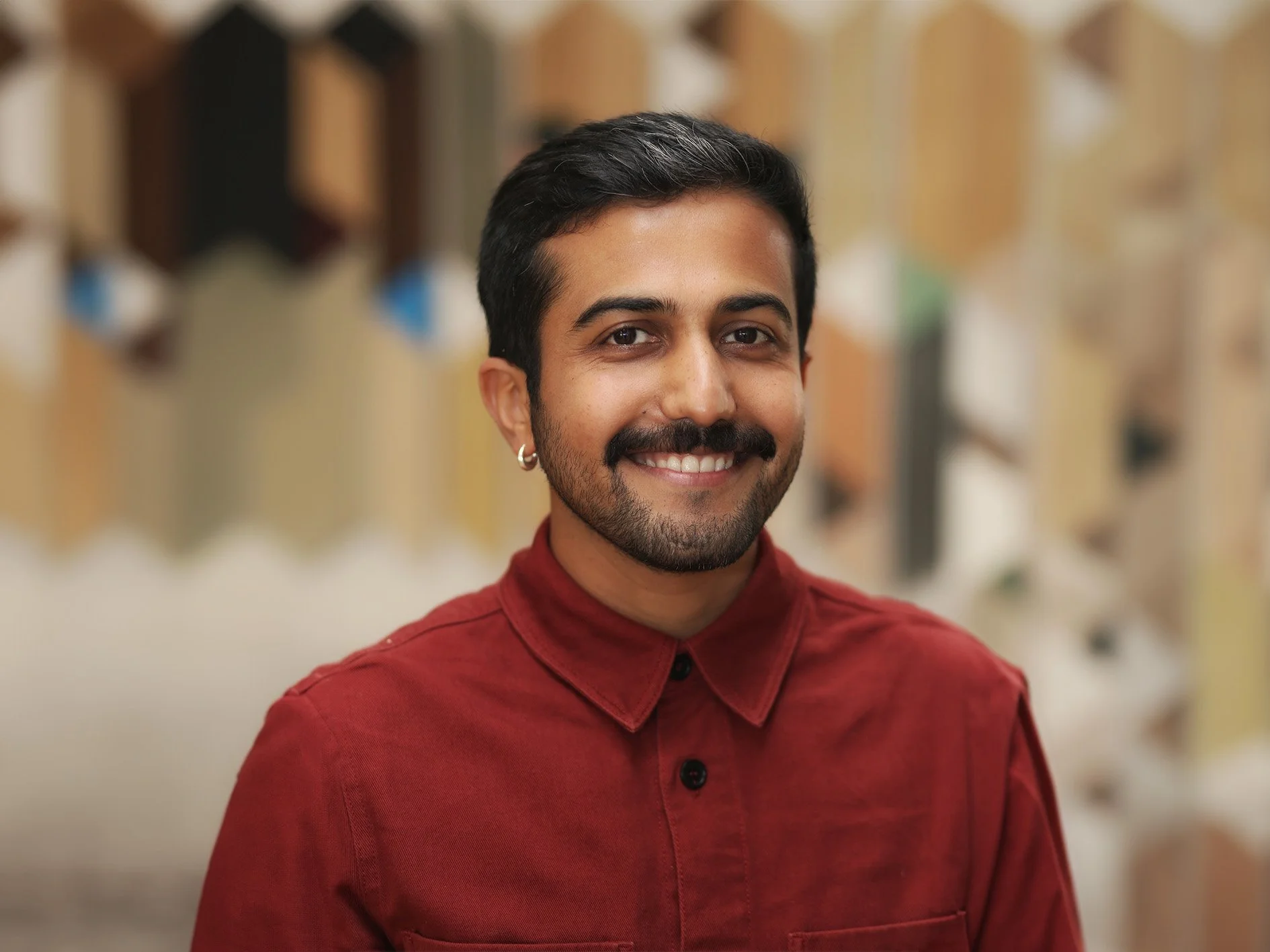 A smiling man with a beard, mustache, and wedding ring earring, wearing a red button-up shirt in front of a blurred background of bookshelves.