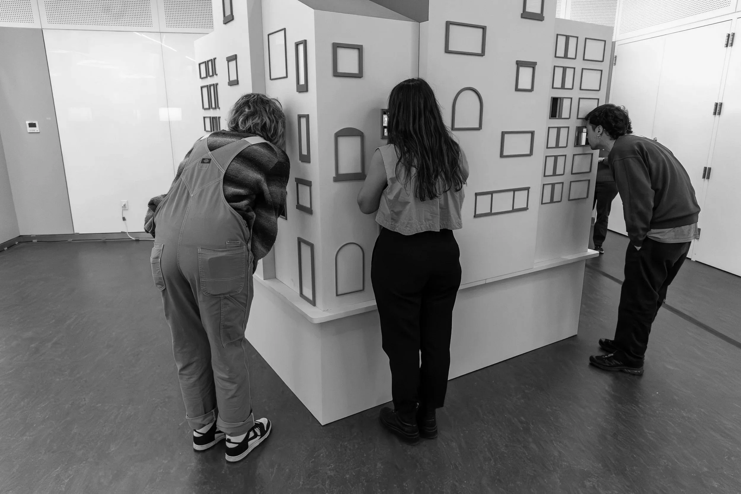 Three people are looking at a display wall with multiple empty picture frames arranged in an asymmetrical pattern.