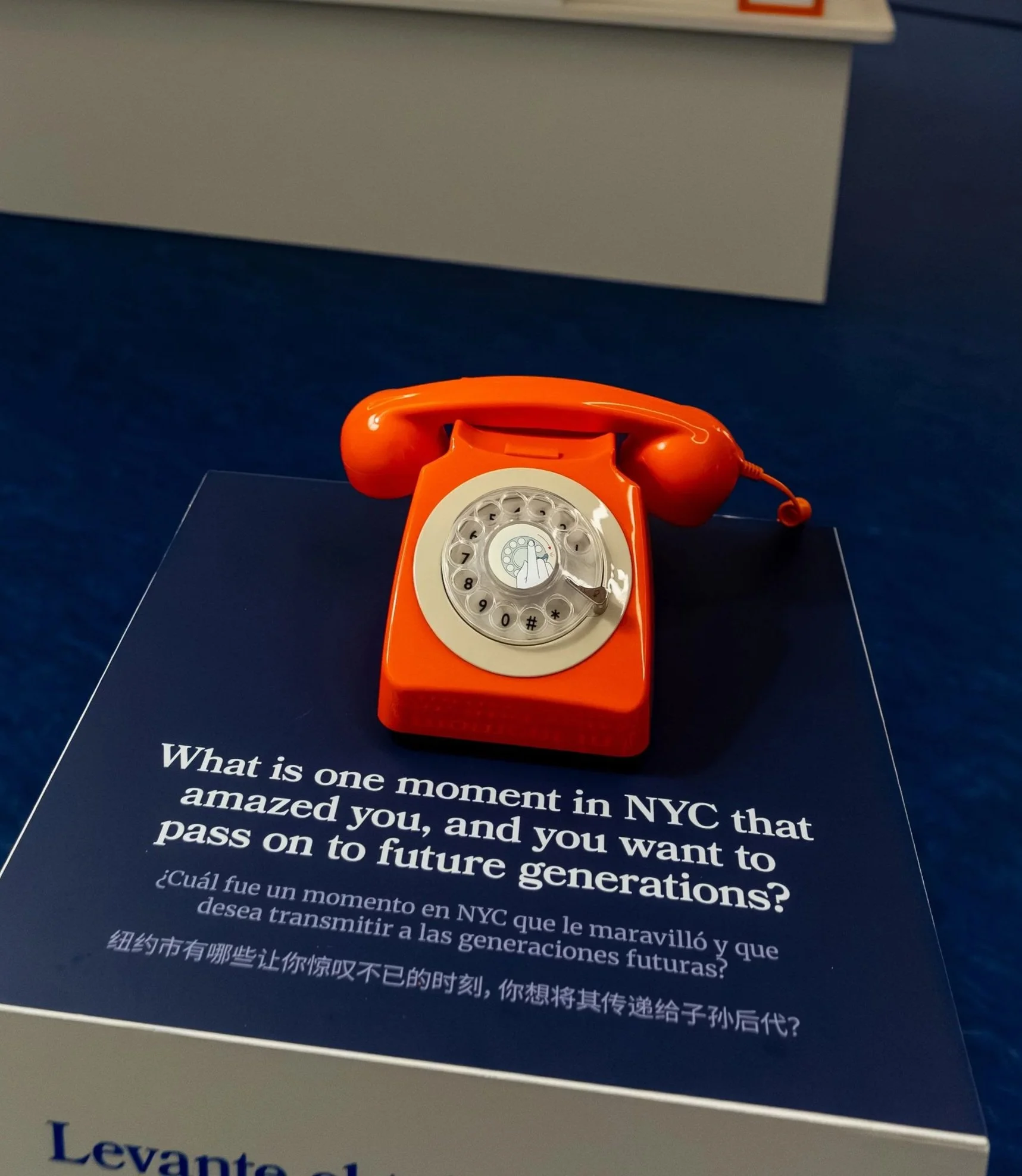 A bright orange rotary dial telephone placed on top of a blue informational display with white text in English and Spanish.