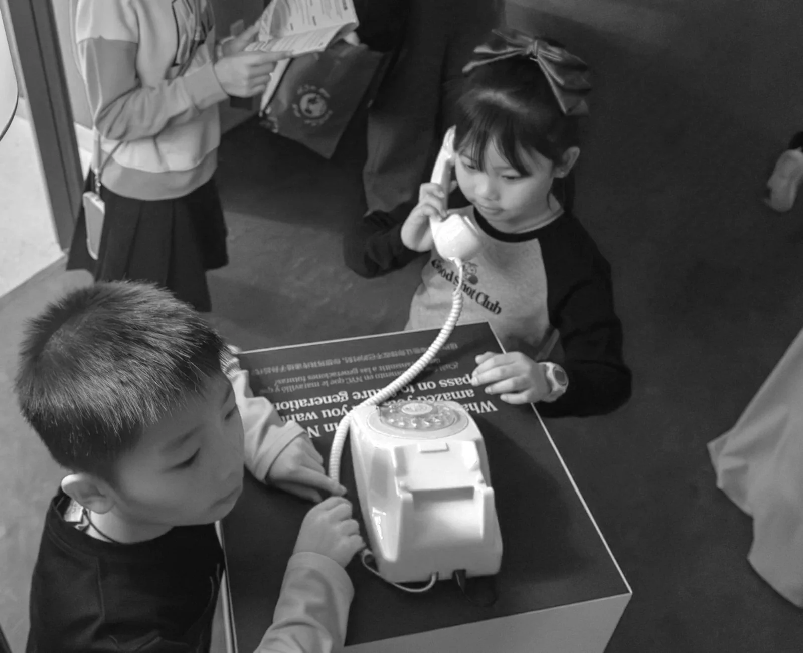Children using a vintage rotary telephone at a desk, with one child speaking into the phone and another watching.