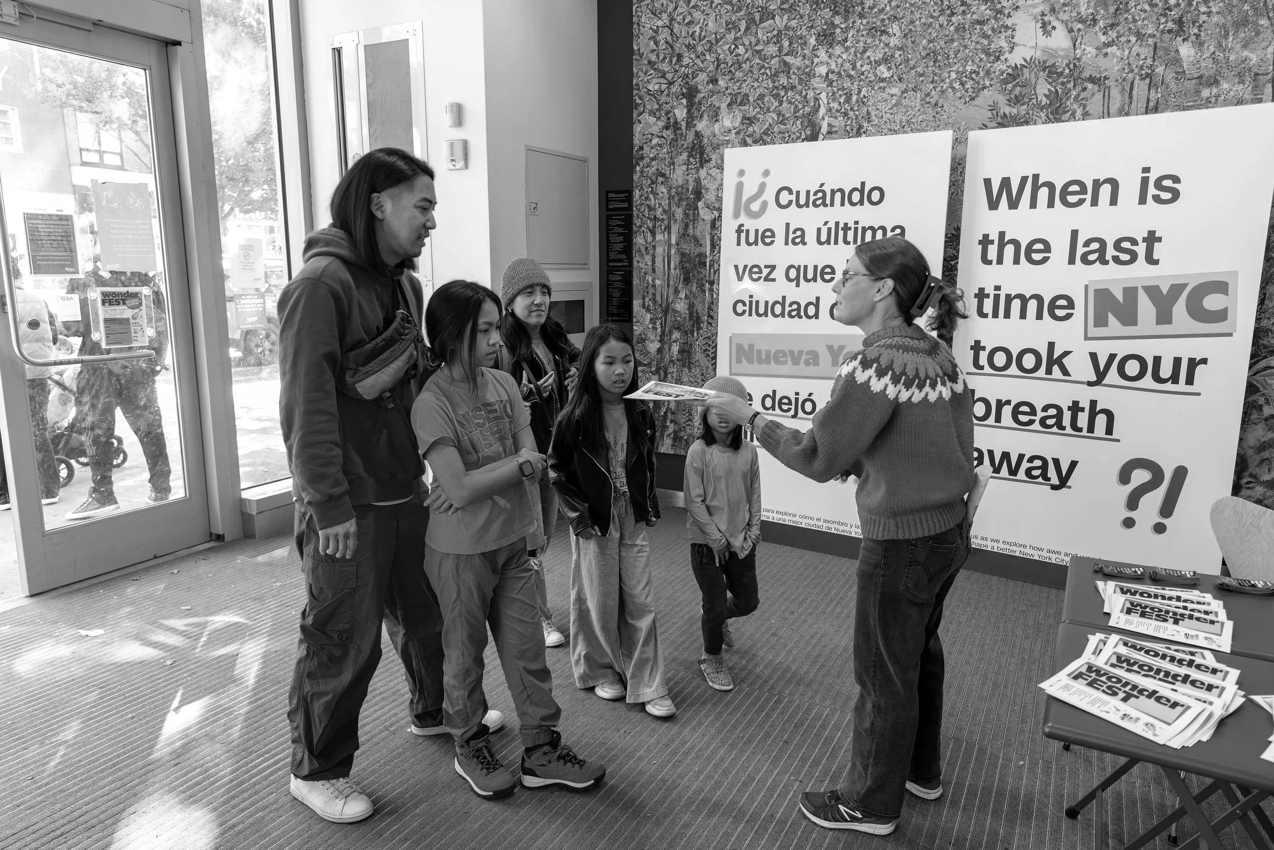 A woman in glasses and a patterned sweater is speaking to a group of four children and one adult at an indoor event. The children are listening attentively. Behind them are large signs with bilingual text about New York City, and a table with pamphlets. The setting appears to be a community or informational center.