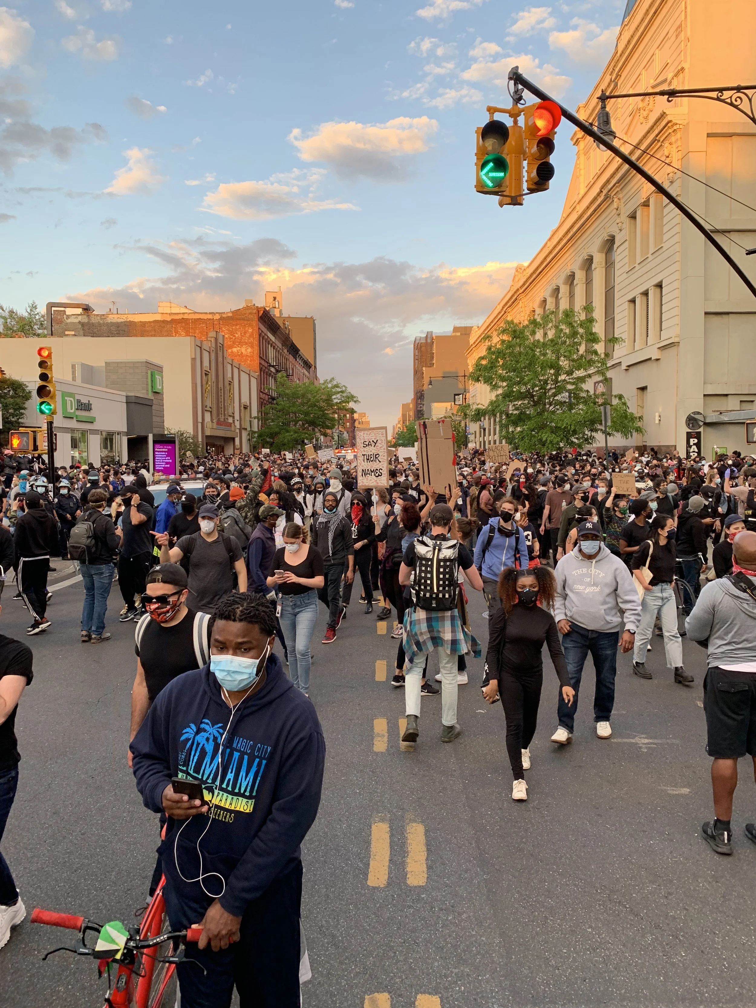 A large crowd of people participating in a protest or demonstration in an urban street, holding signs and wearing masks, with traffic lights visible overhead and city buildings on either side.