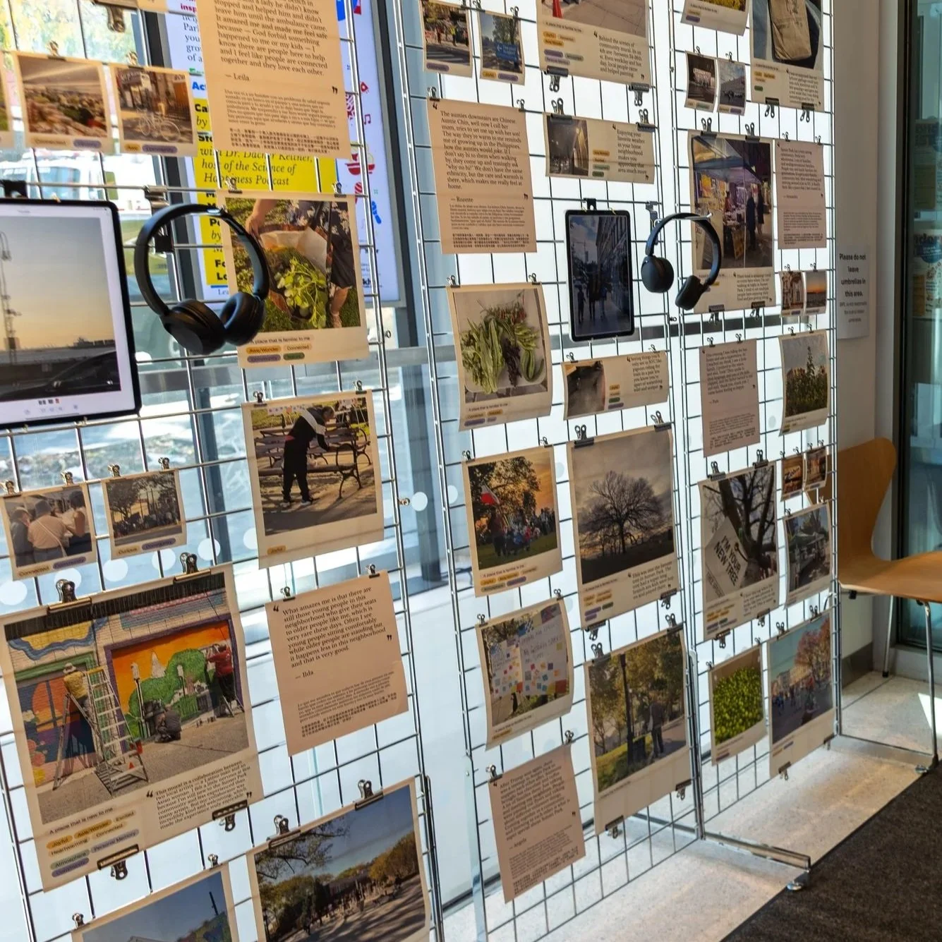 An indoor display of wire grid panels holds many photographs, informational posters, and a pair of black headphones. The background shows large windows revealing parked cars outside.