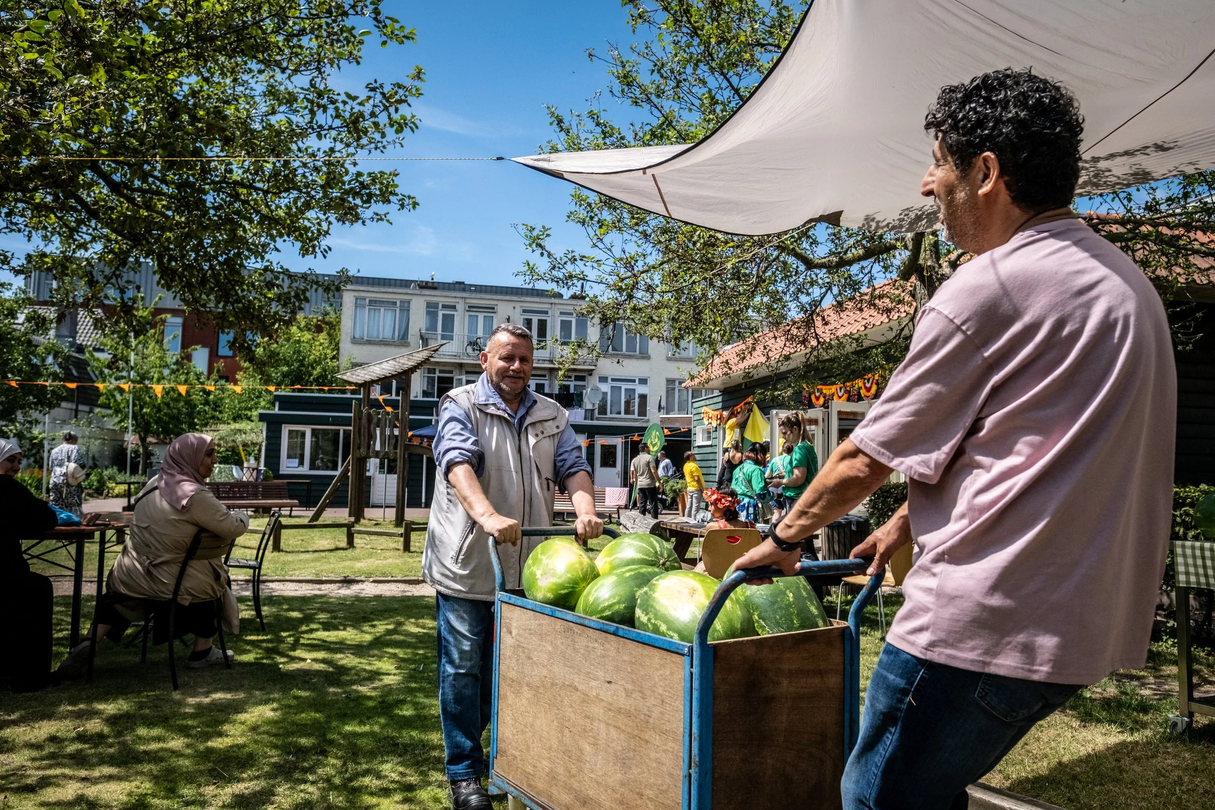 Two men are pushing a cart with watermelons at an outdoor market or festival. In the background, people are sitting, eating, and walking around under trees and umbrellas. There are colorful decorations and buildings, with a clear blue sky.