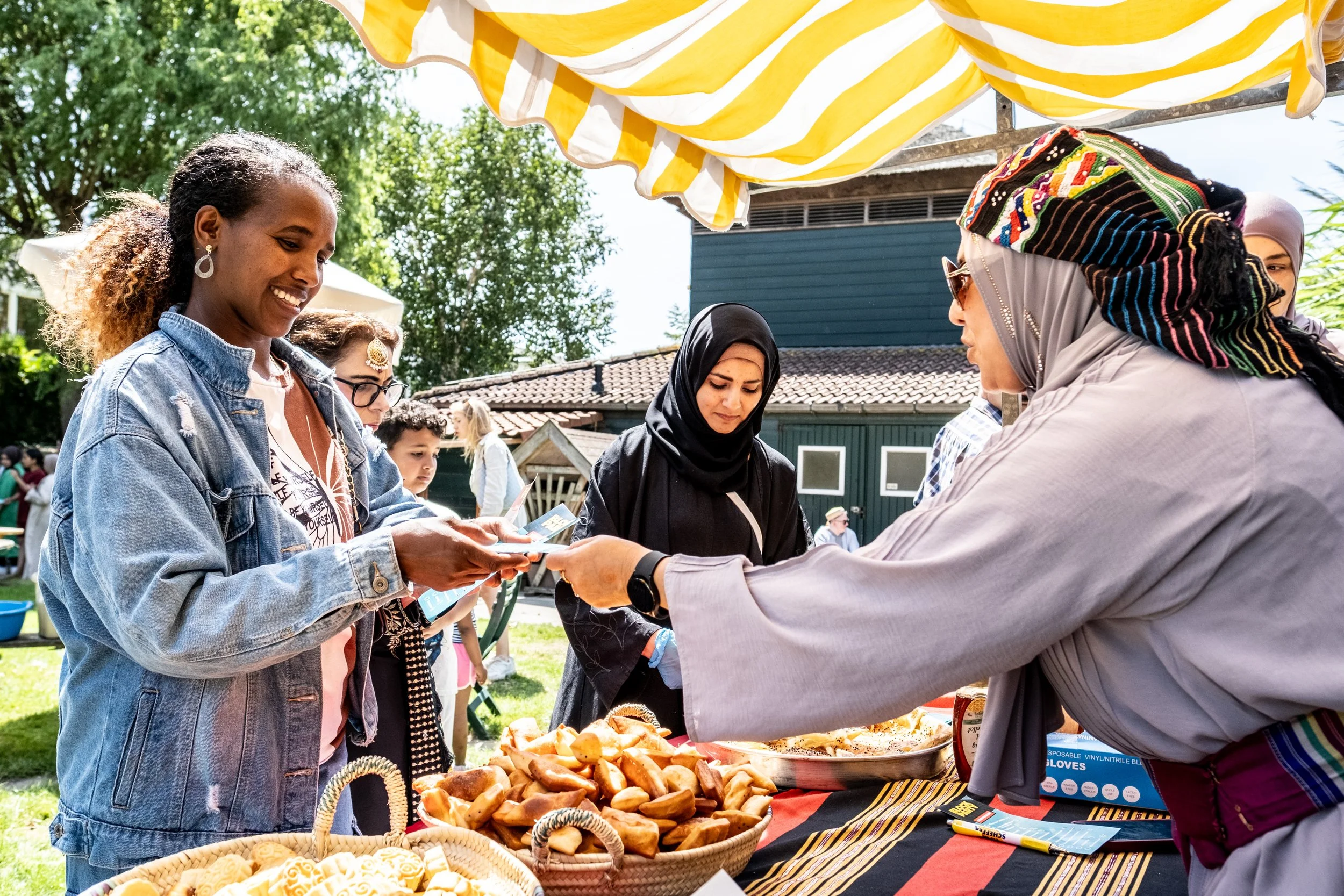 A woman in a black hijab and a woman in a colorful headscarf exchange money or a card at a food stall with baked goods during an outdoor event on a sunny day.