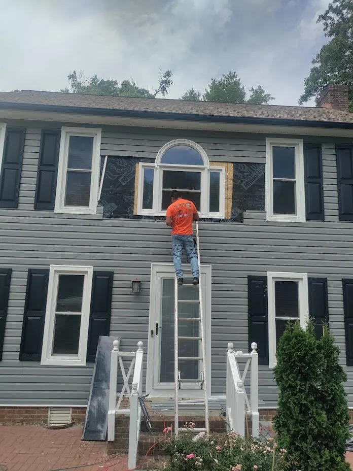 An employee safely works on a ladder on a second floor window, creating custom fitted new siding for a semi circle window. 