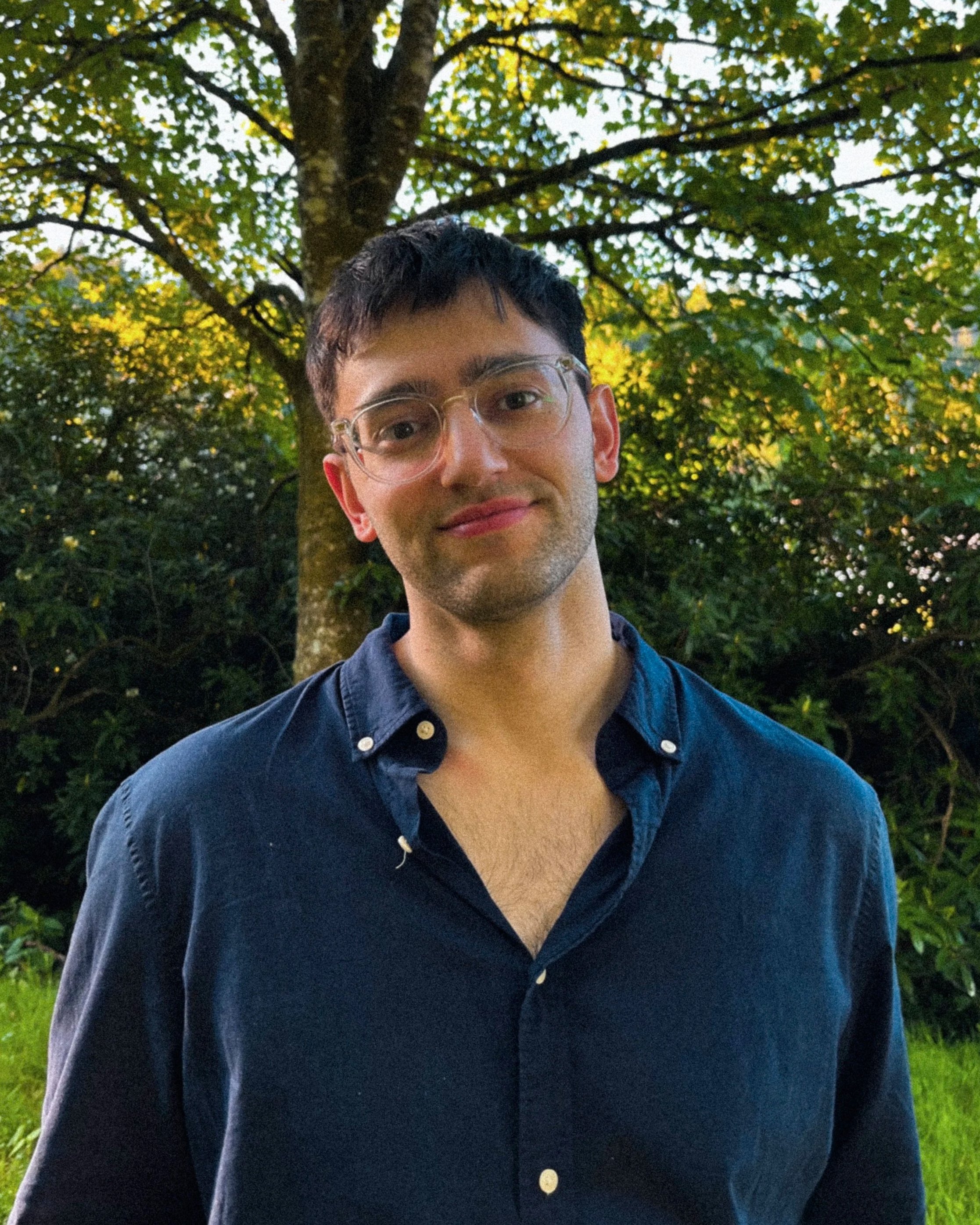 A man with glasses and dark hair is standing outdoors in front of a large tree with green leaves, wearing a dark button-up shirt, during twilight.