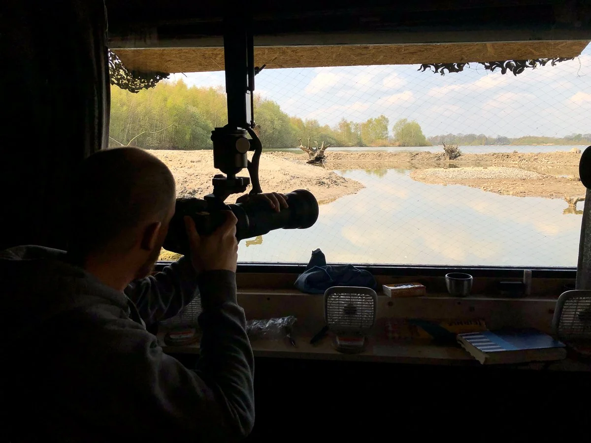Mensch mit Fernglas und Kamera in einem Tarnzelt beim Beobachten der Natur an einem Flussufer mit Wasser und Bäumen im Hintergrund.