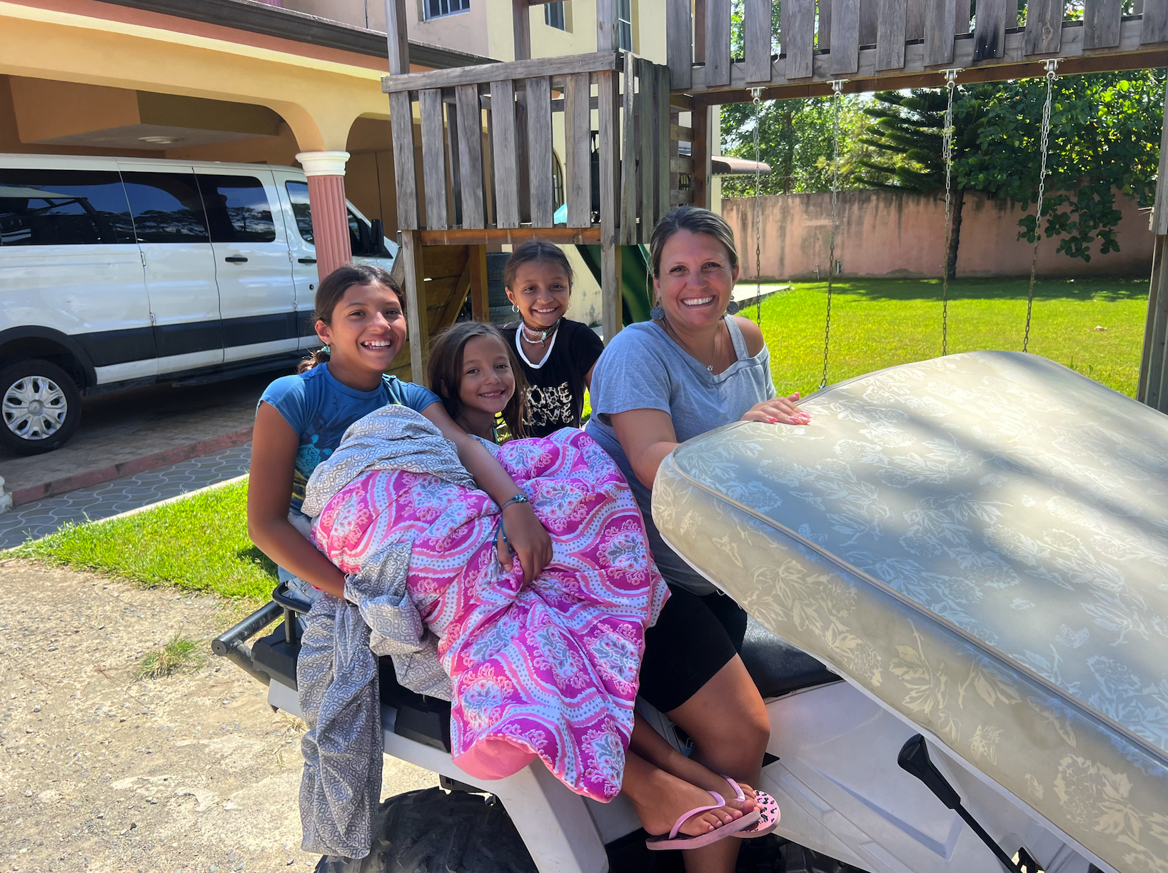 A woman and three young girls sitting on a golf cart outdoors. The woman is smiling and the girls are sitting with blankets. A house, a fence, and a grassy yard are in the background.