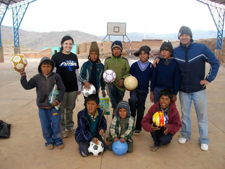 Group of children and two adults on an outdoor basketball court, holding soccer balls, with mountains in the background.