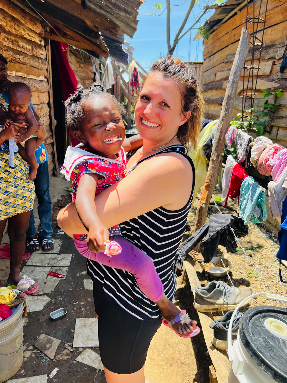 A woman and a young girl happily hugging outside in a rural area with wooden structures. The woman is wearing a striped tank top and black shorts, smiling at the camera. The girl is dressed in colorful clothes, holding a lollipop, and smiling. Laundr